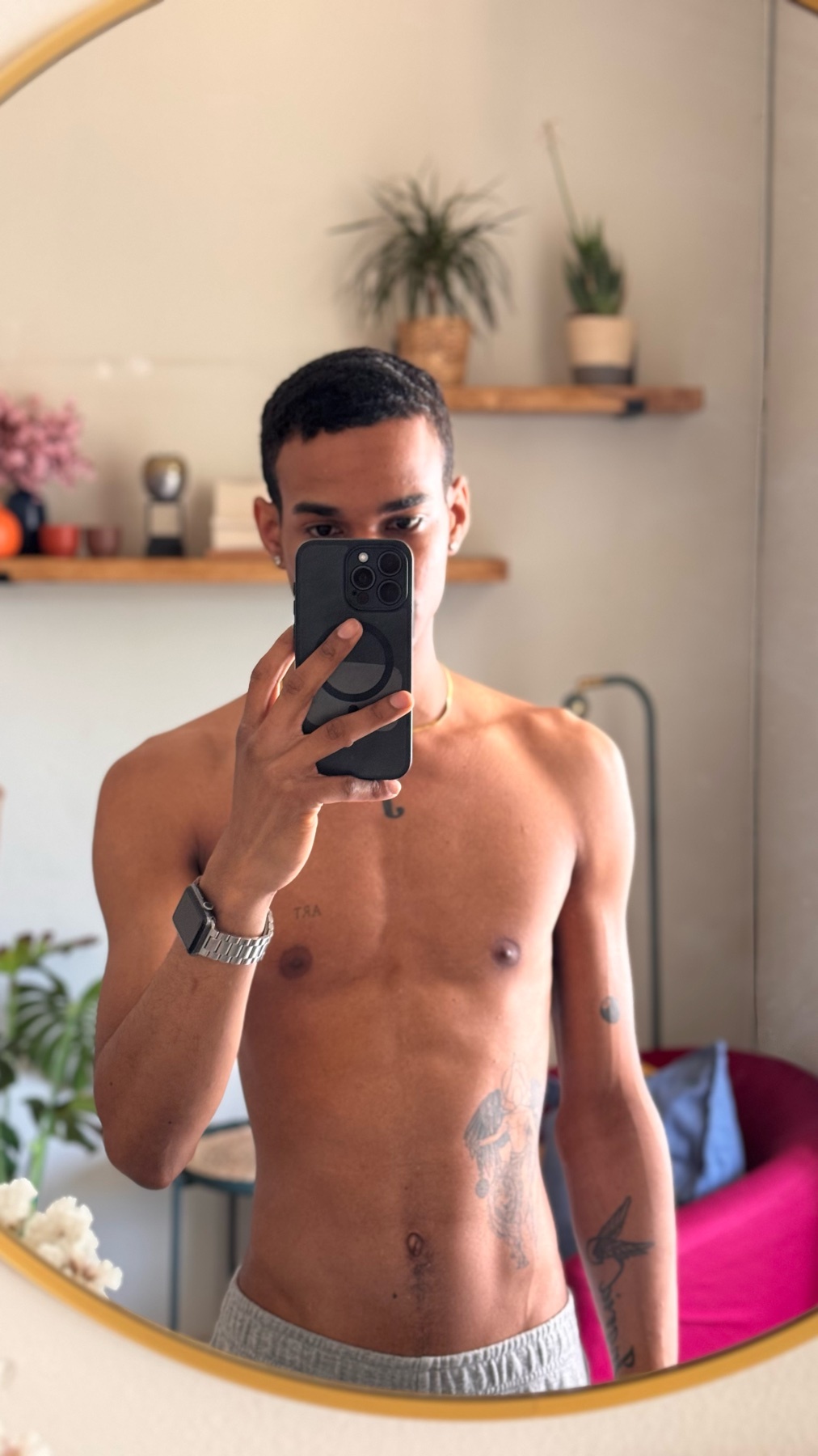 A shirtless man with tattoos takes a mirror selfie in his home. He is wearing grey sweatpants and a watch.