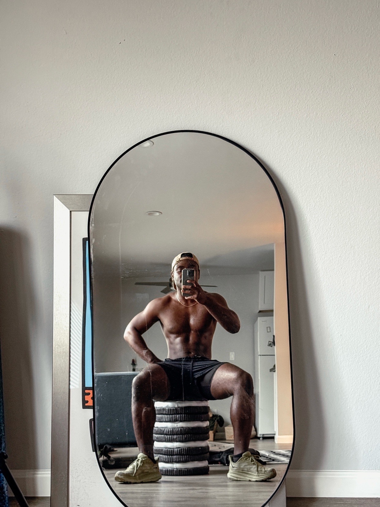 A shirtless man poses in front of a mirror, showcasing his muscular physique. He is wearing shorts and a cap, with weights and gym equipment visible in the background.