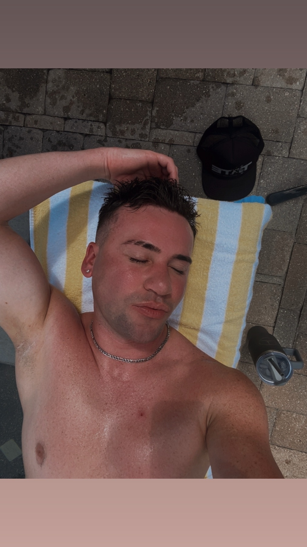 A man relaxes poolside, wearing a necklace and a hat nearby. He appears to be sweating and has tan lines.