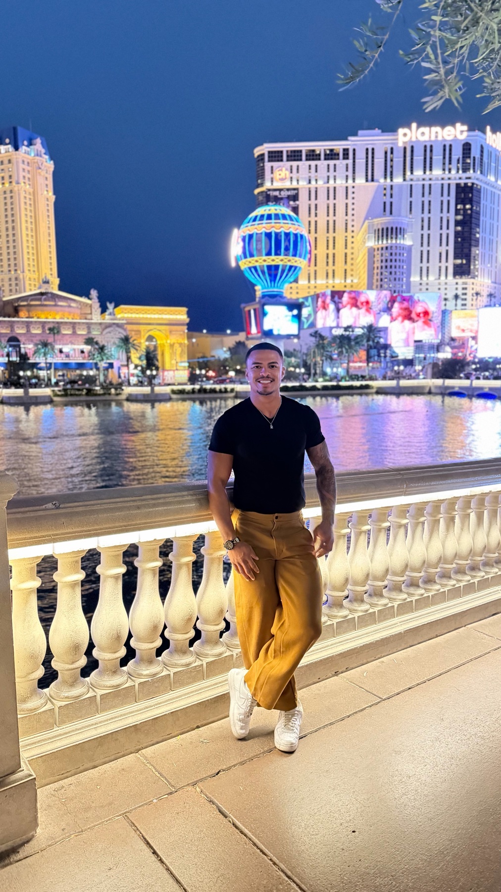 A man poses for a photo at night in front of the Las Vegas strip. The cityscape is lit up with bright lights and a body of water is visible in the background.