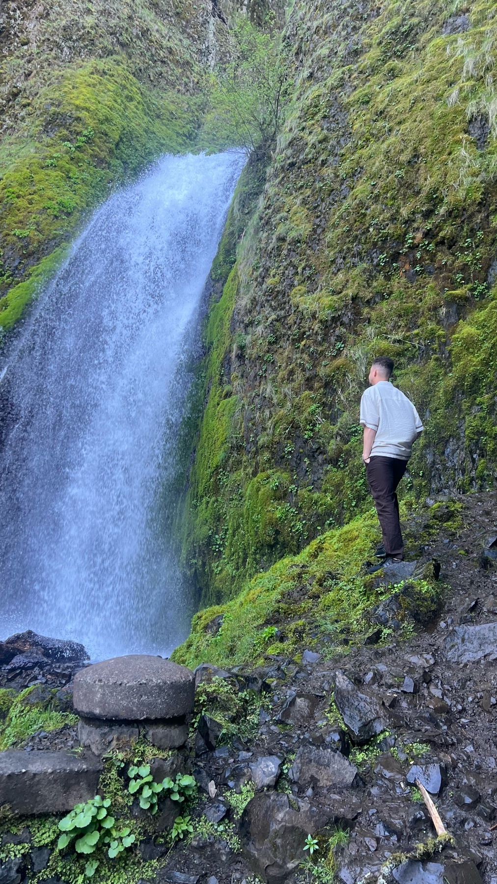 A man stands on a rocky path, looking up at a tall, cascading waterfall surrounded by lush, moss-covered cliffs.