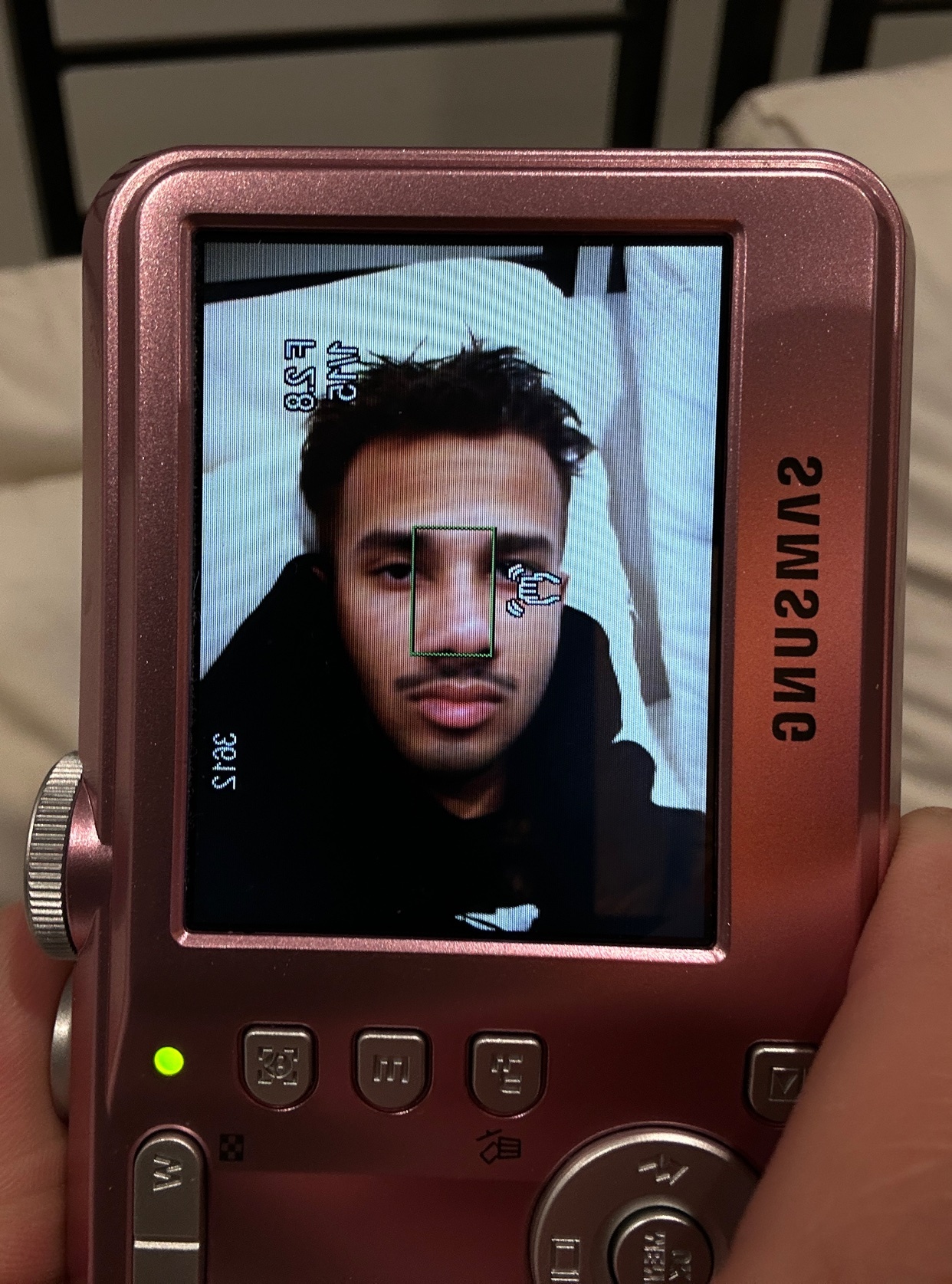 A man takes a selfie with a pink digital camera. The camera screen shows a close-up of his face with a mustache and stubble.