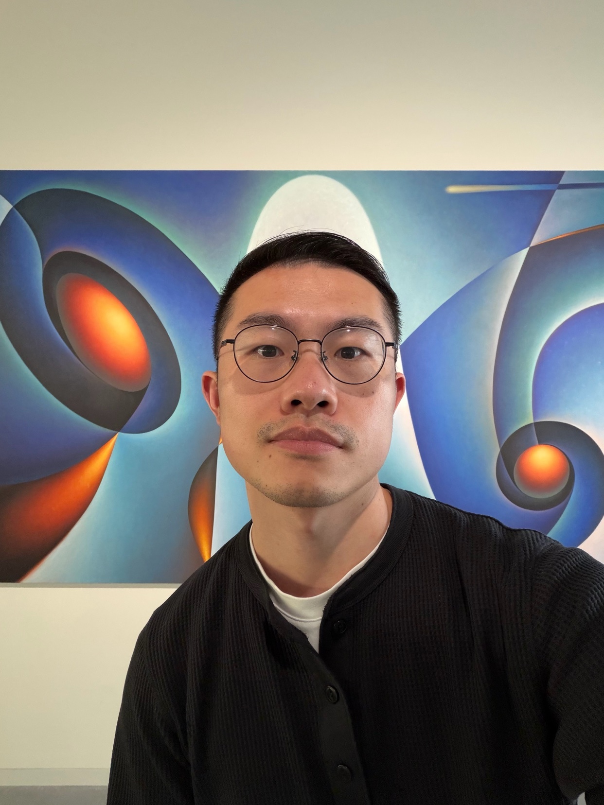 A man with glasses and a white hat takes a selfie in front of a vibrant, abstract painting featuring swirling blue and orange shapes.
