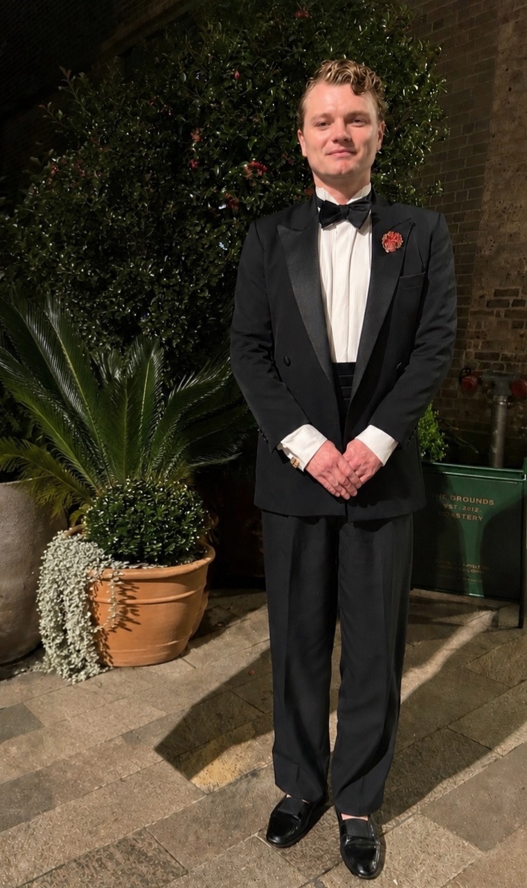 A man stands in a formal black tuxedo with a white shirt and black bow tie, accessorized with a poppy pin on his lapel. He is dressed for a black tie event and stands outdoors with greenery in the background.