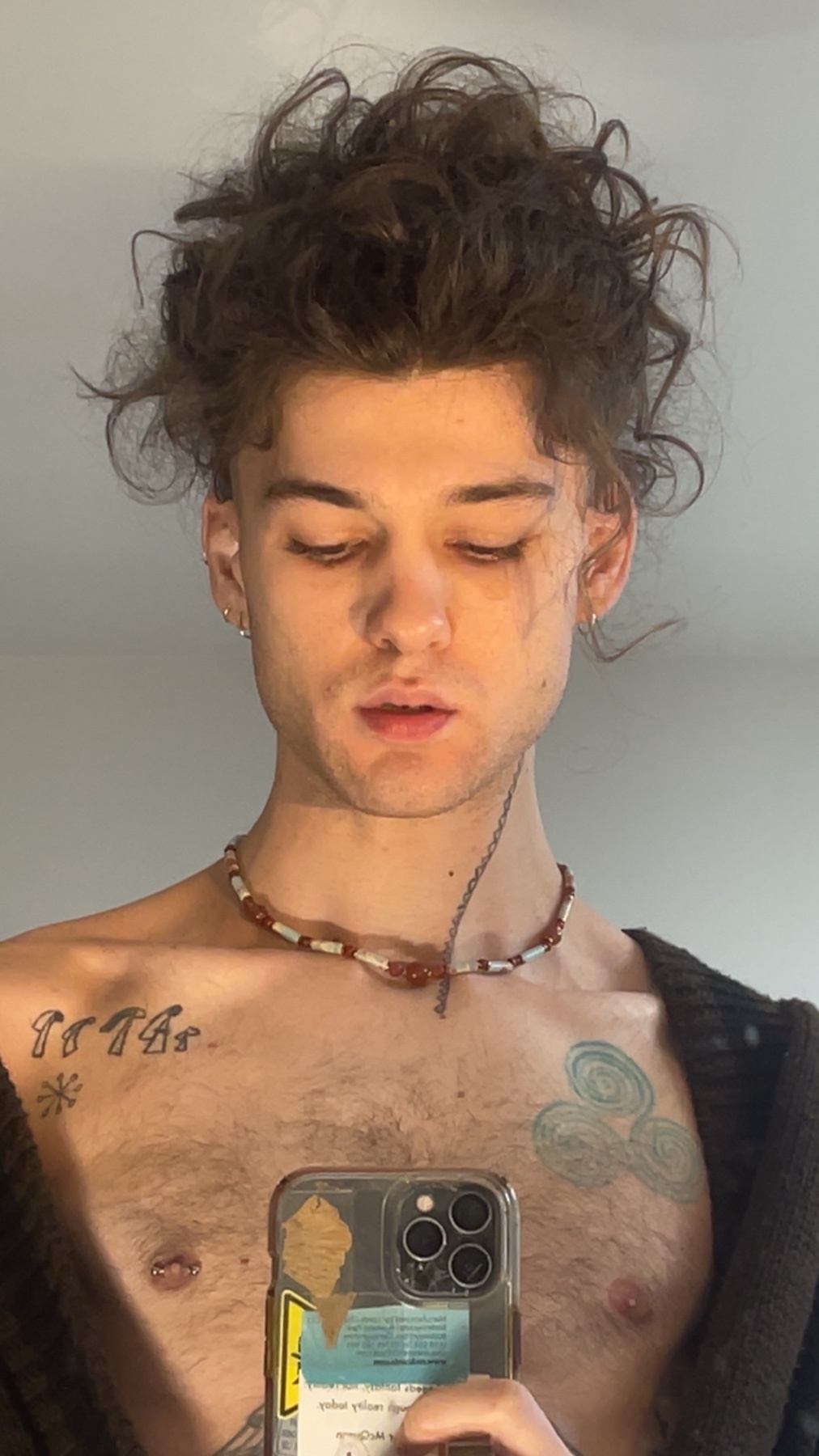 A man takes a mirror selfie, shirtless, showcasing his tattoos and chest hair. His curly hair is styled up, and he wears a necklace.