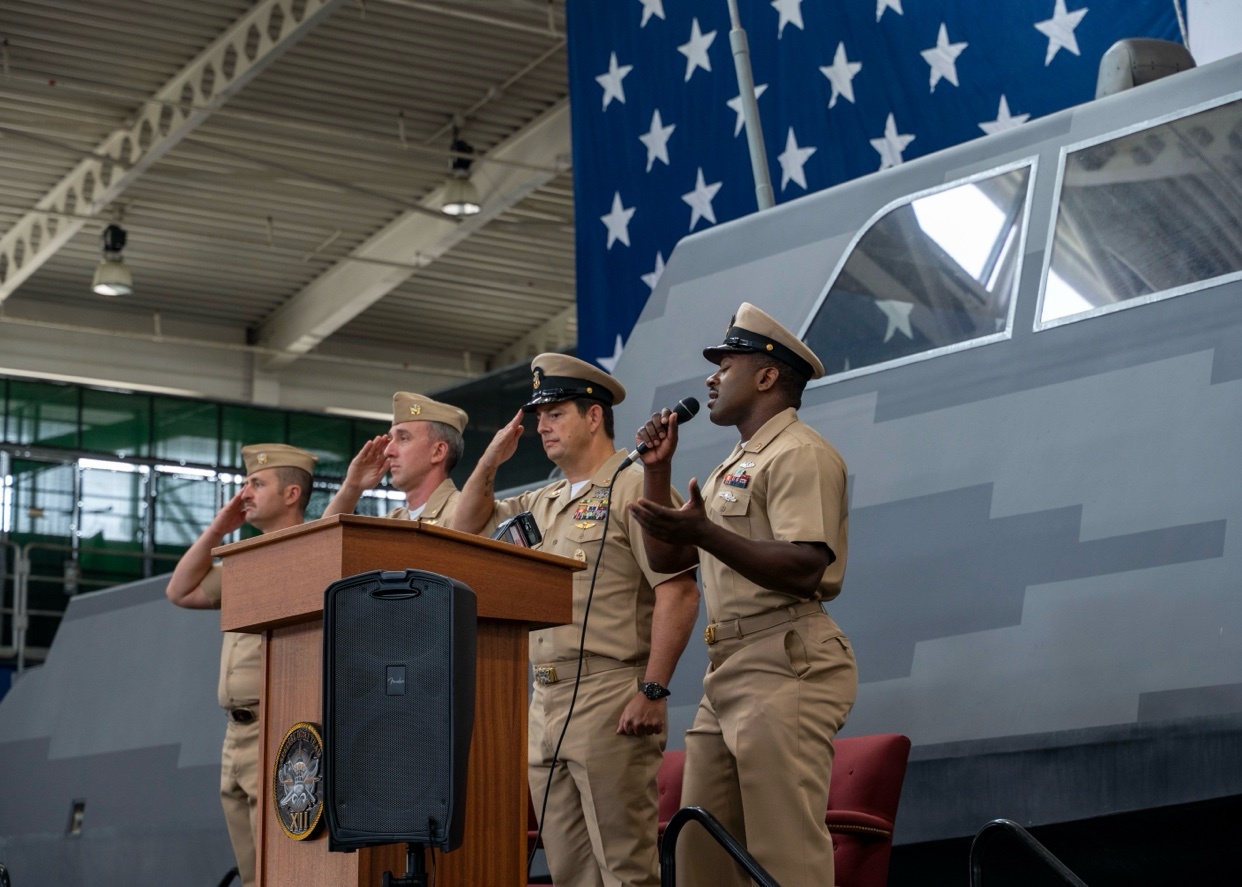Four men in navy uniforms stand at a podium with a large American flag in the background. One man is speaking into a microphone while the other three salute.