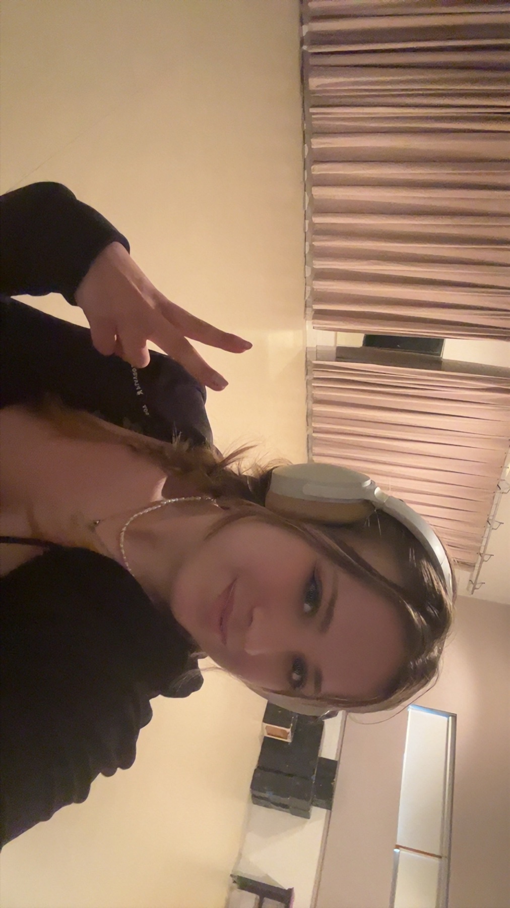 A young woman with long, braided blonde hair takes a selfie while wearing headphones and making a peace sign with her fingers. She is wearing a necklace and a dark top.