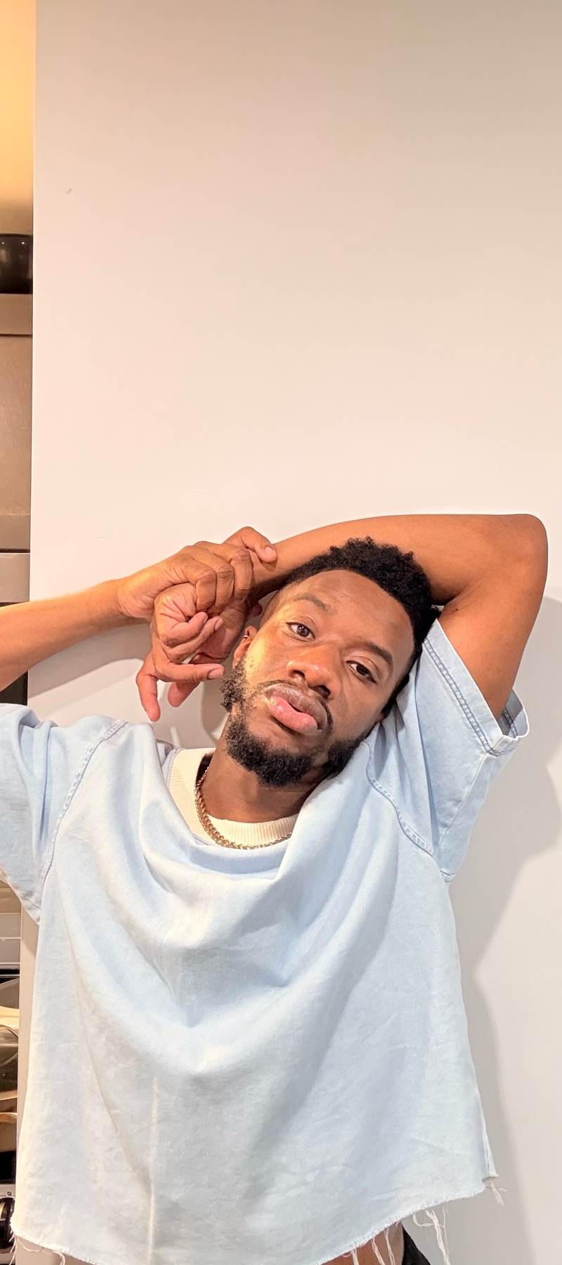 A man poses for a photo, leaning against a wall. He is wearing a light blue t-shirt.