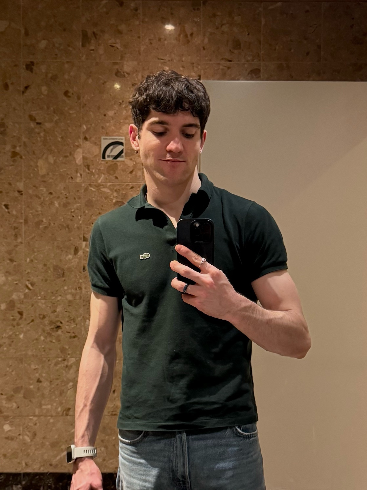 A man takes a selfie in a dark green polo shirt and jeans, with curly dark hair and a watch on his left wrist.