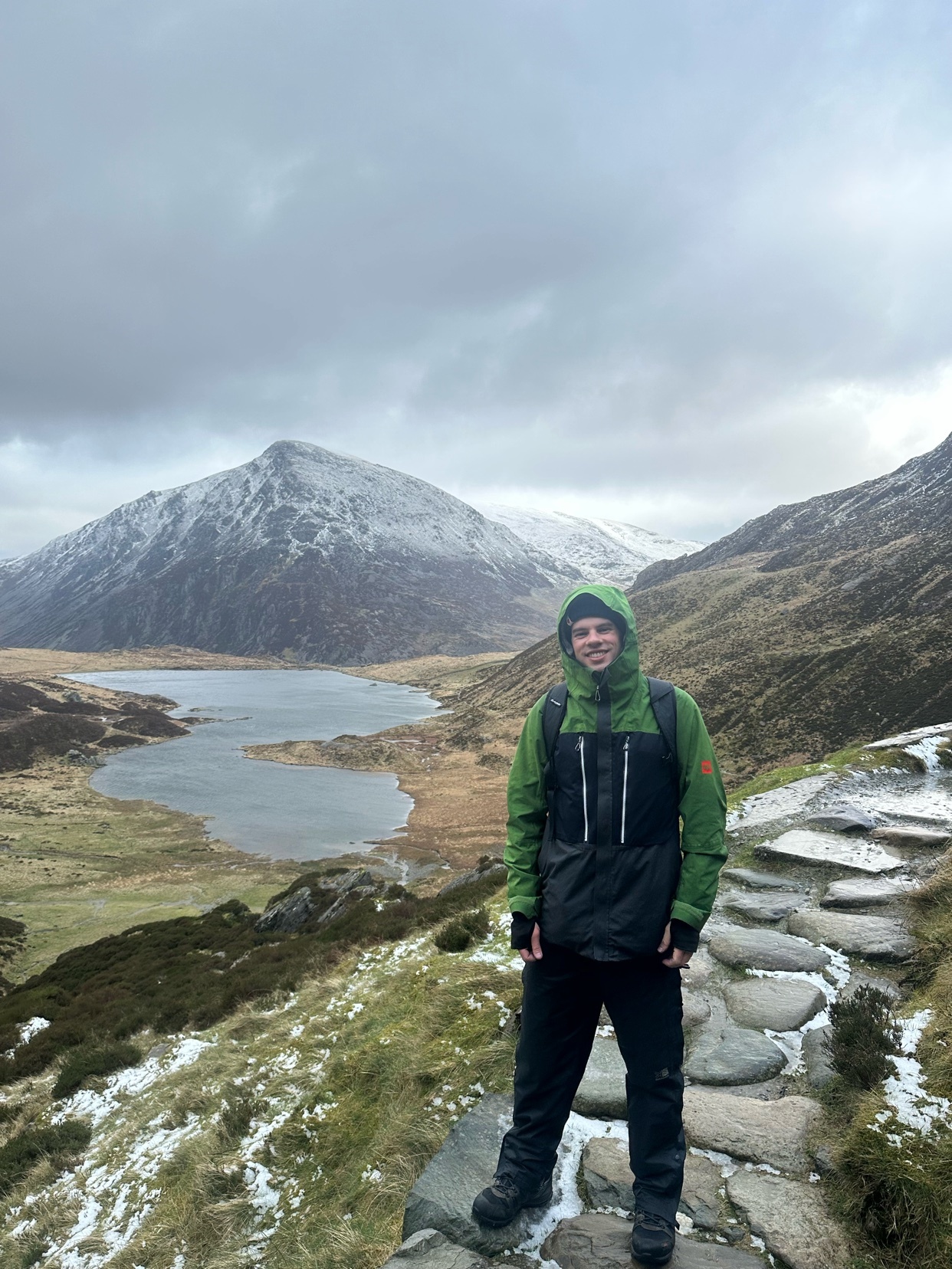 A man stands on a stone path in a mountainous landscape with snow on the ground and peaks. He is wearing a green hooded jacket and black pants, with a backpack on.