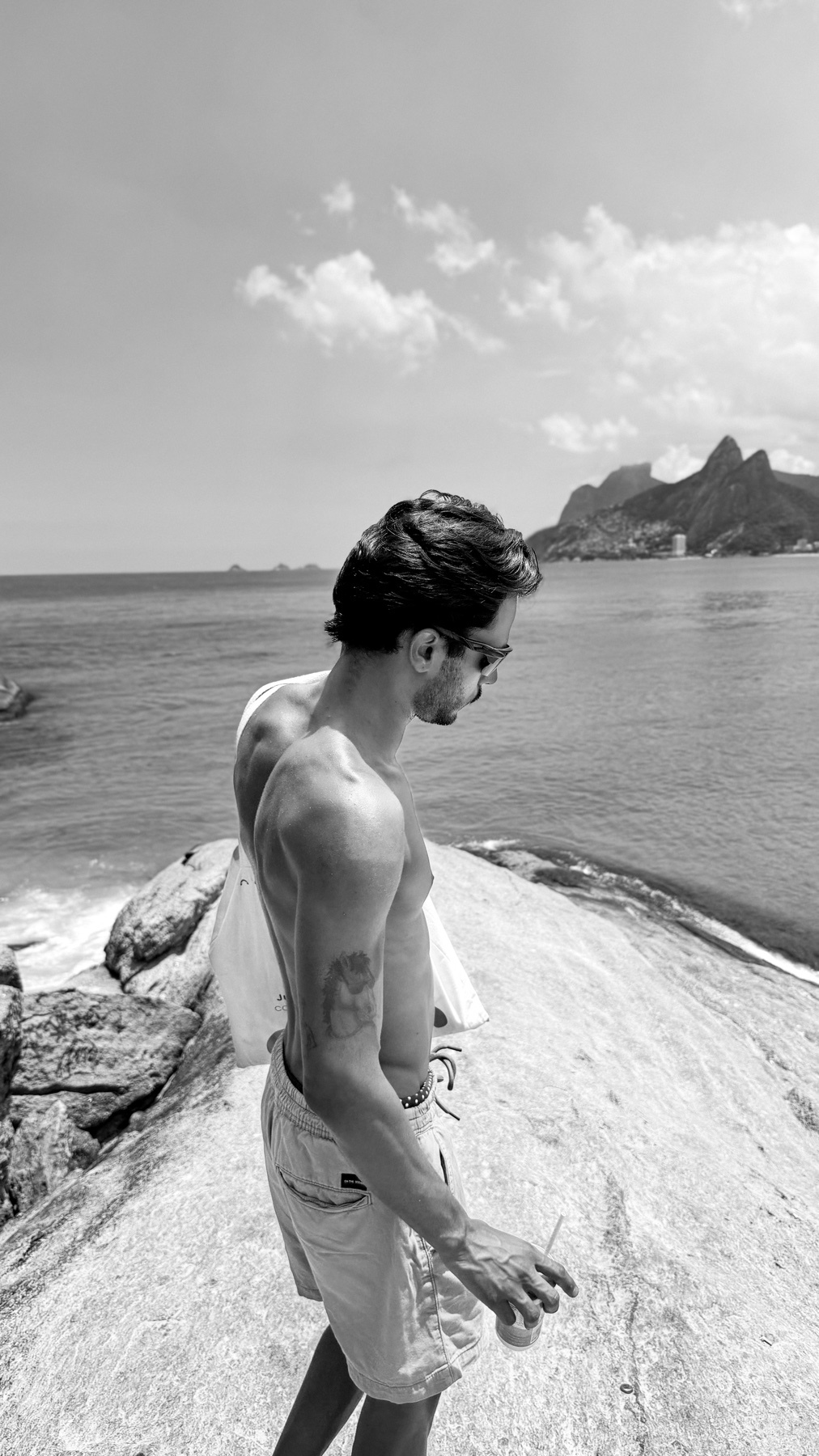A black and white, medium shot of a man standing on a rocky outcrop overlooking the ocean. He is shirtless, wearing shorts and sunglasses, with a tattoo on his arm. Mountains are visible in the background.