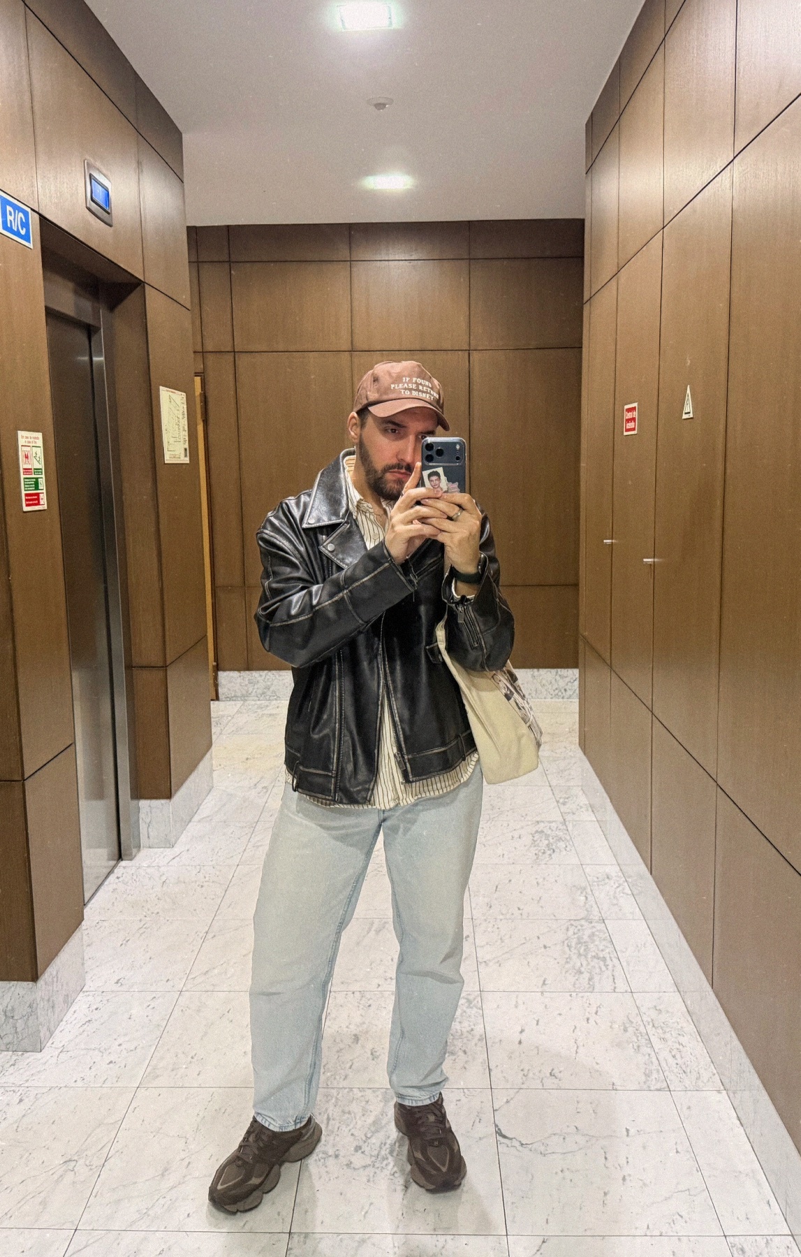 A man takes a mirror selfie in an elevator. He is wearing a leather jacket, jeans, and a cap.