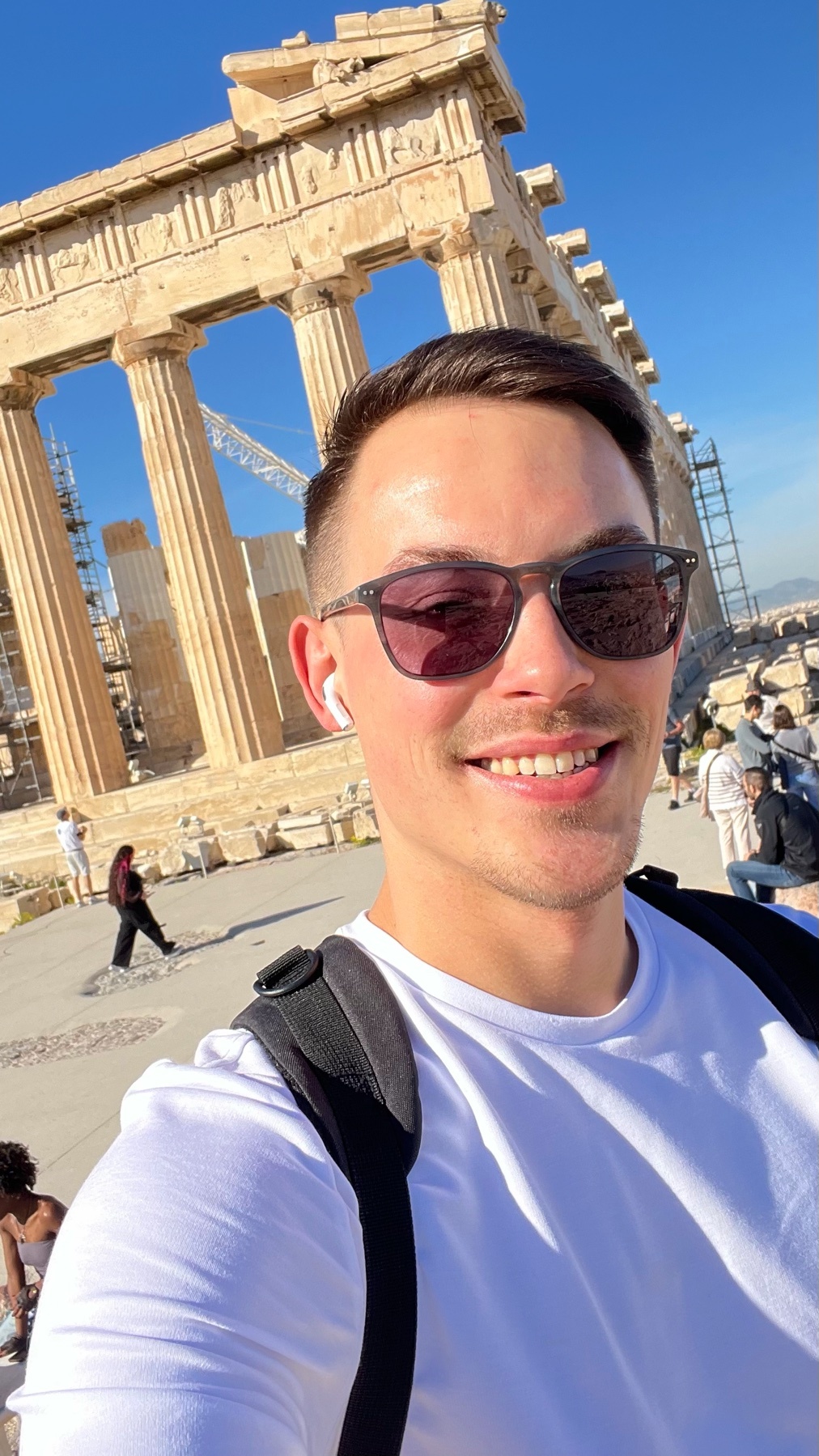 A man takes a selfie in front of the Parthenon in Athens, Greece. He is smiling and wearing sunglasses and a white t-shirt.