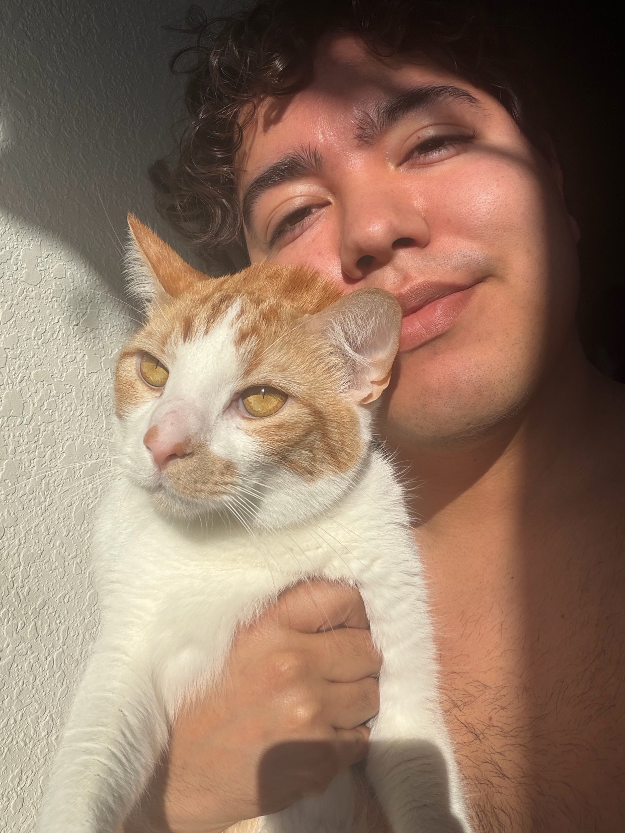 A man with curly dark hair takes a selfie with his orange and white cat. The man is shirtless and holding the cat close to his face.