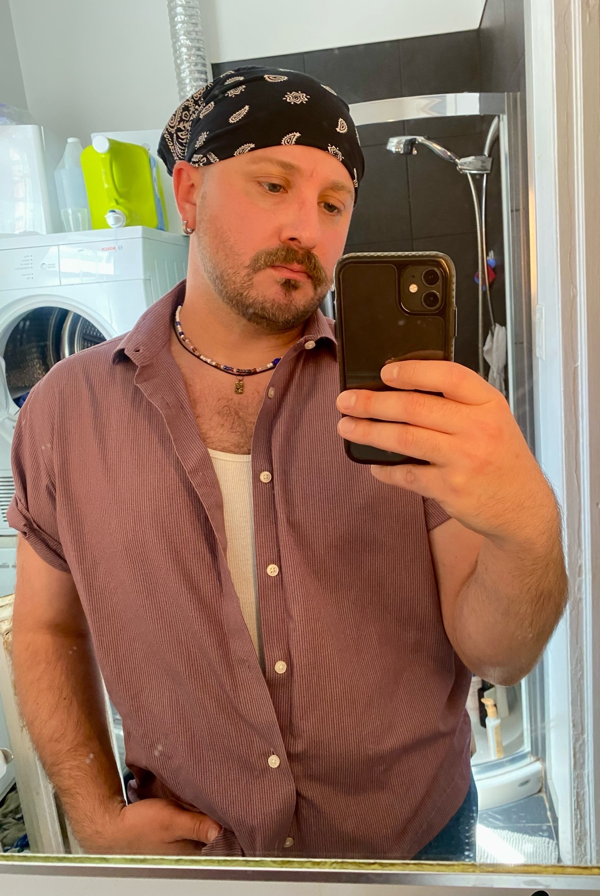 A man with a bandana and mustache takes a selfie in a bathroom. He is wearing a button-up shirt over a tank top and a necklace.