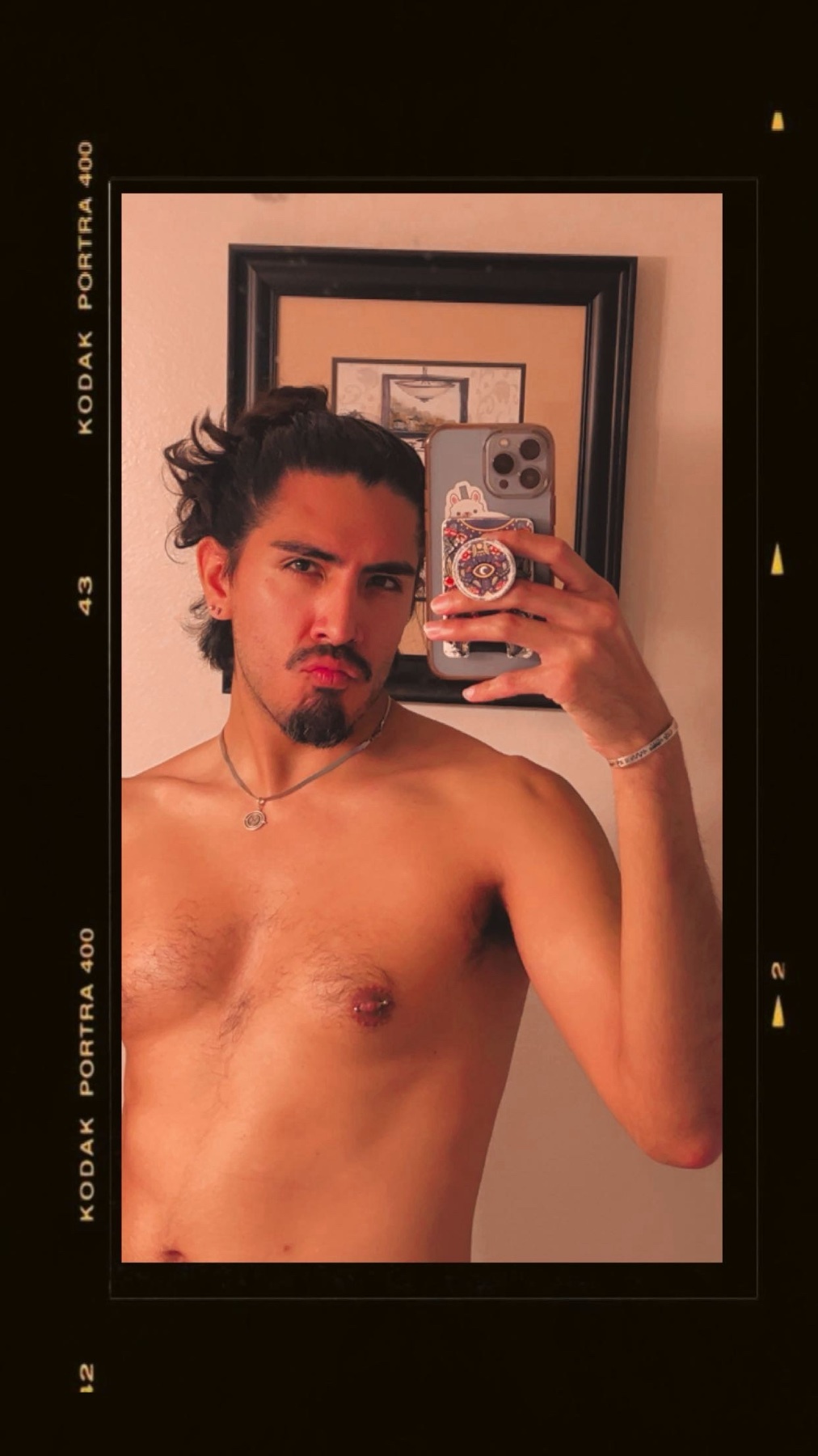 A man with long, dark, curly hair pulled back into a bun takes a selfie in a vintage film style. He is shirtless and has a mustache and goatee.