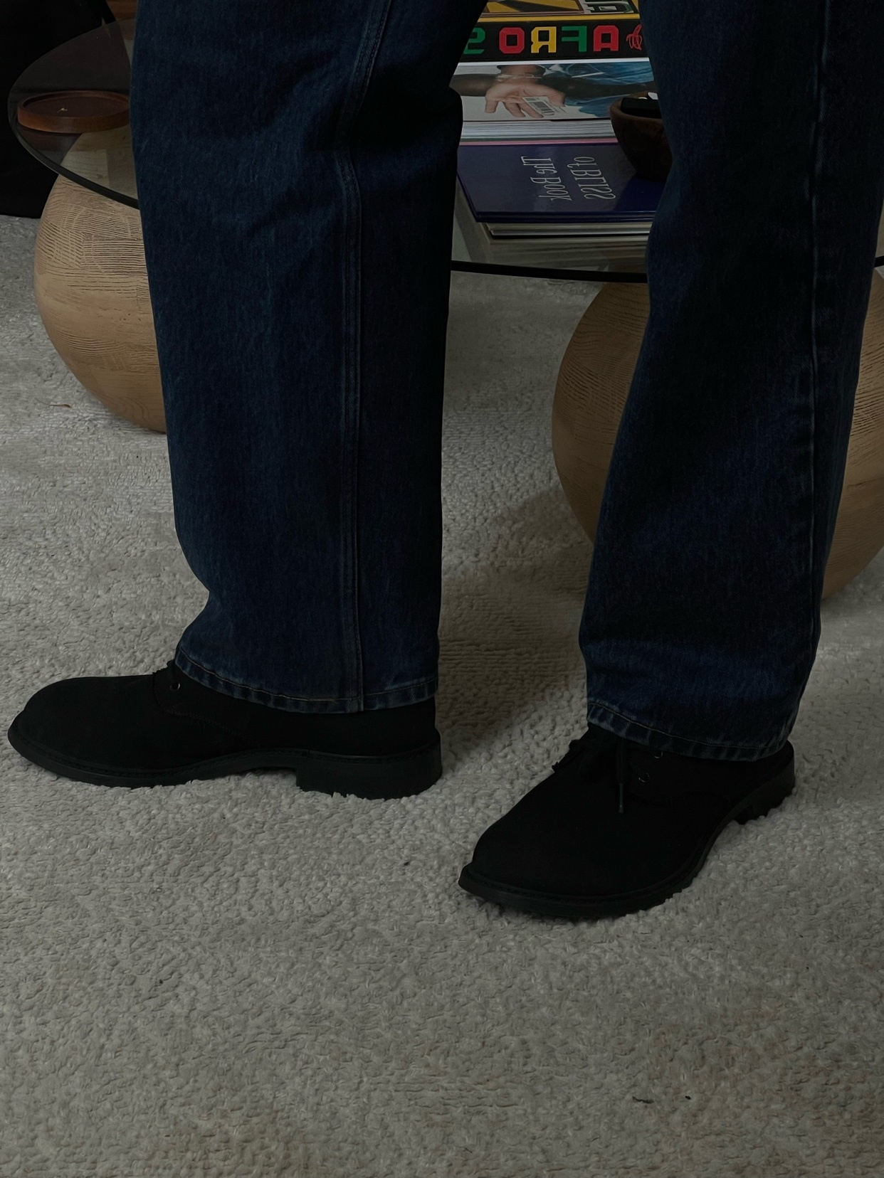A person's legs are shown from the knees down, wearing dark wash jeans and black boots. The boots are a simple, modern style.