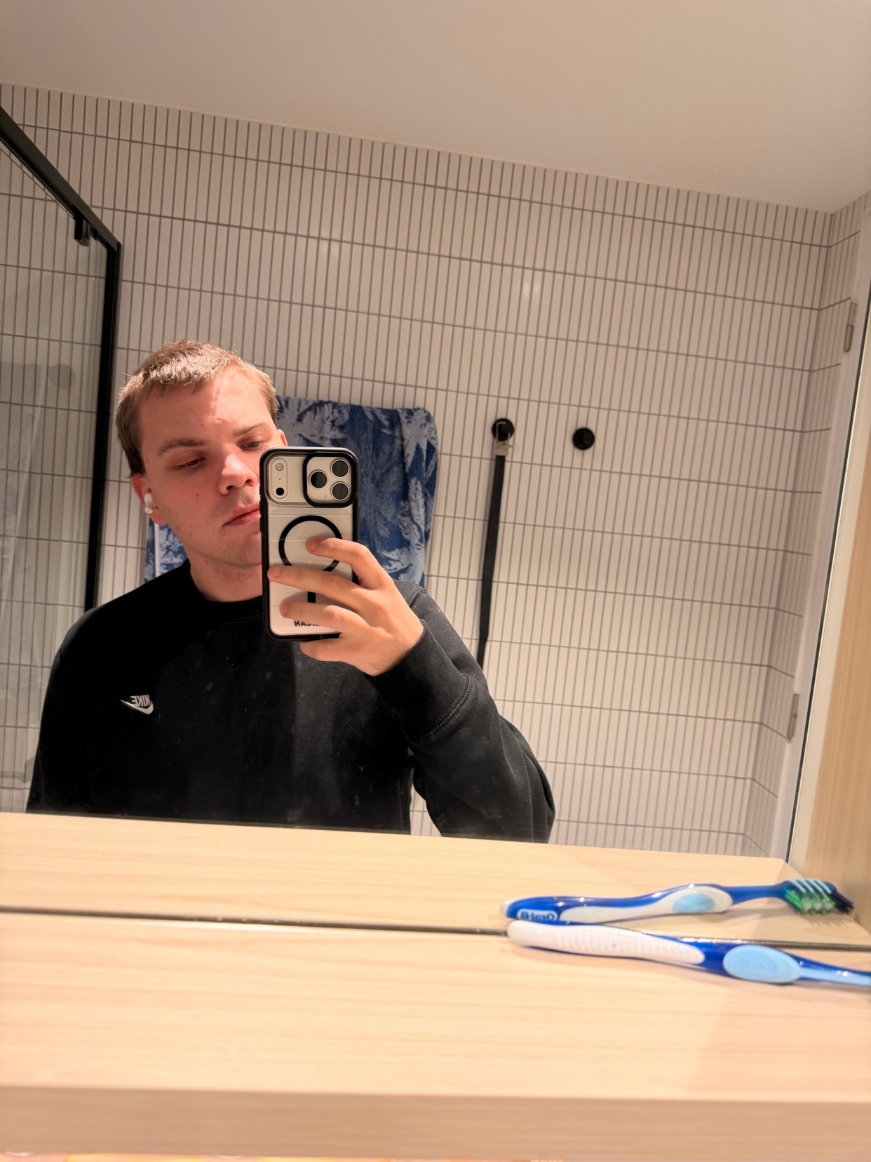 A young man takes a mirror selfie in a bathroom. He is wearing a black sweatshirt with a Nike logo and wireless earbuds.