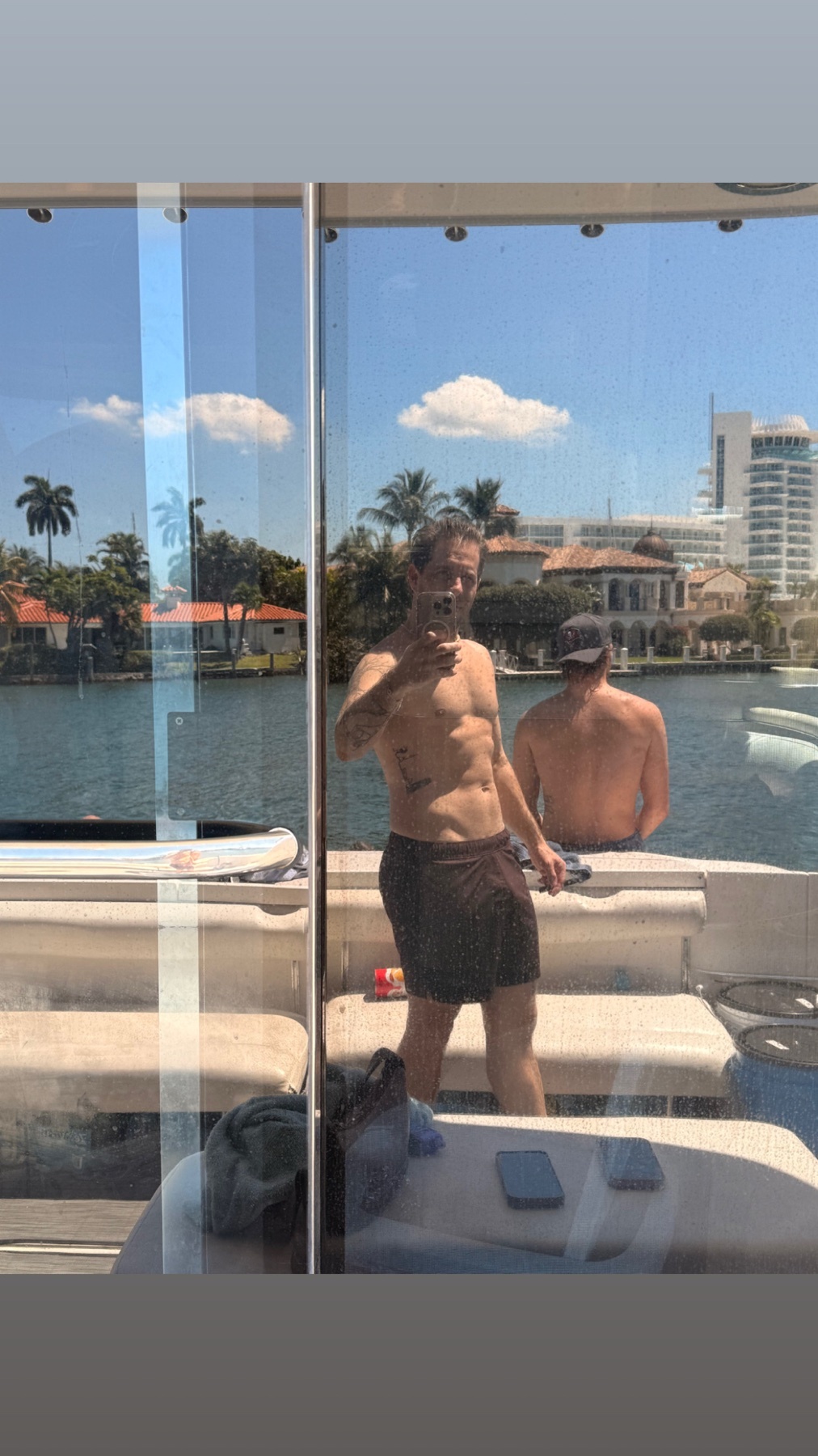 A man with tattoos and a muscular physique takes a selfie on a yacht while another man stands behind him. The background shows a sunny day with palm trees and buildings.