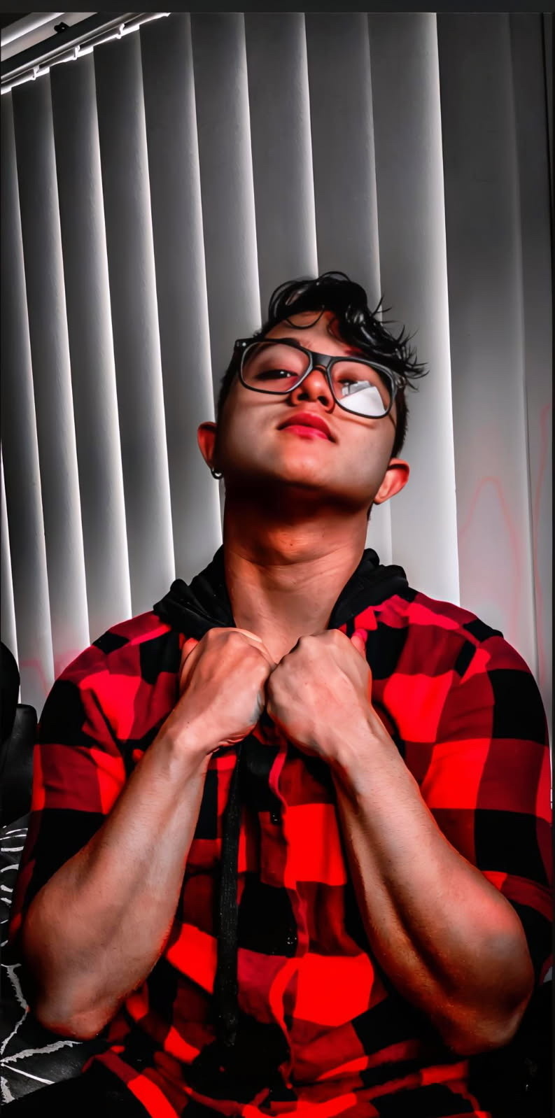 A man with dark, styled hair and glasses poses in a red and black plaid flannel shirt with a hood. His arms are crossed in front of his chest, and his muscles are visible.