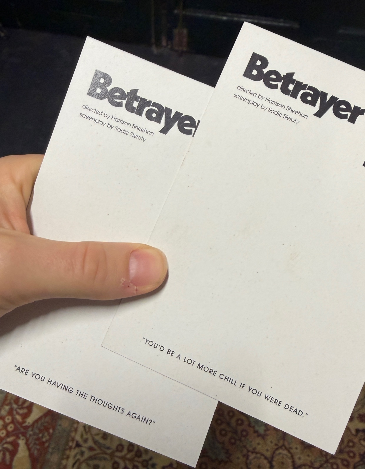 A person holds two tickets for the play "Betrayer." The tickets are white with black text, featuring quotes from the play.