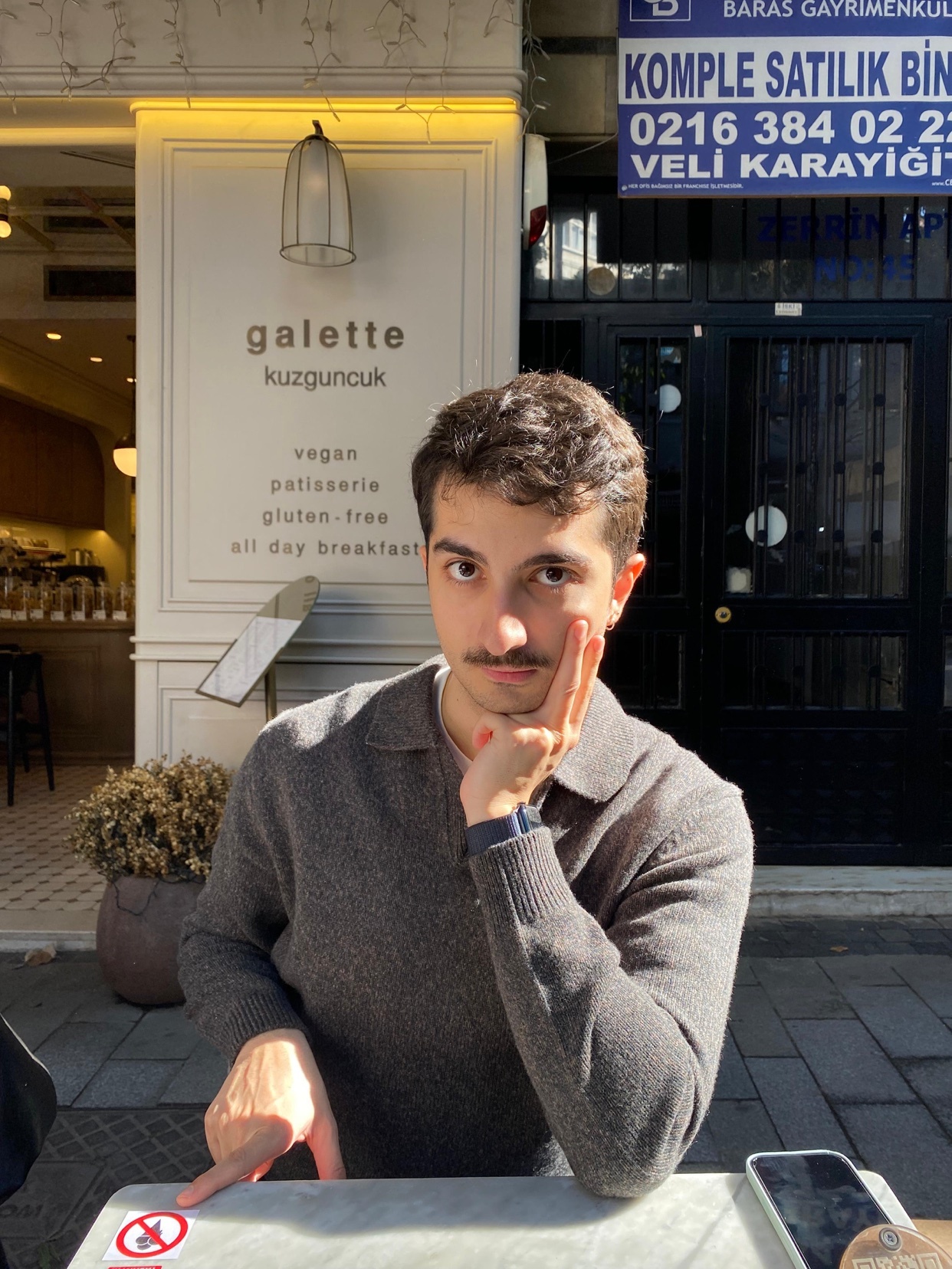 A man poses at a cafe table, resting his hand on his cheek. The cafe, "galette," offers vegan and gluten-free options.