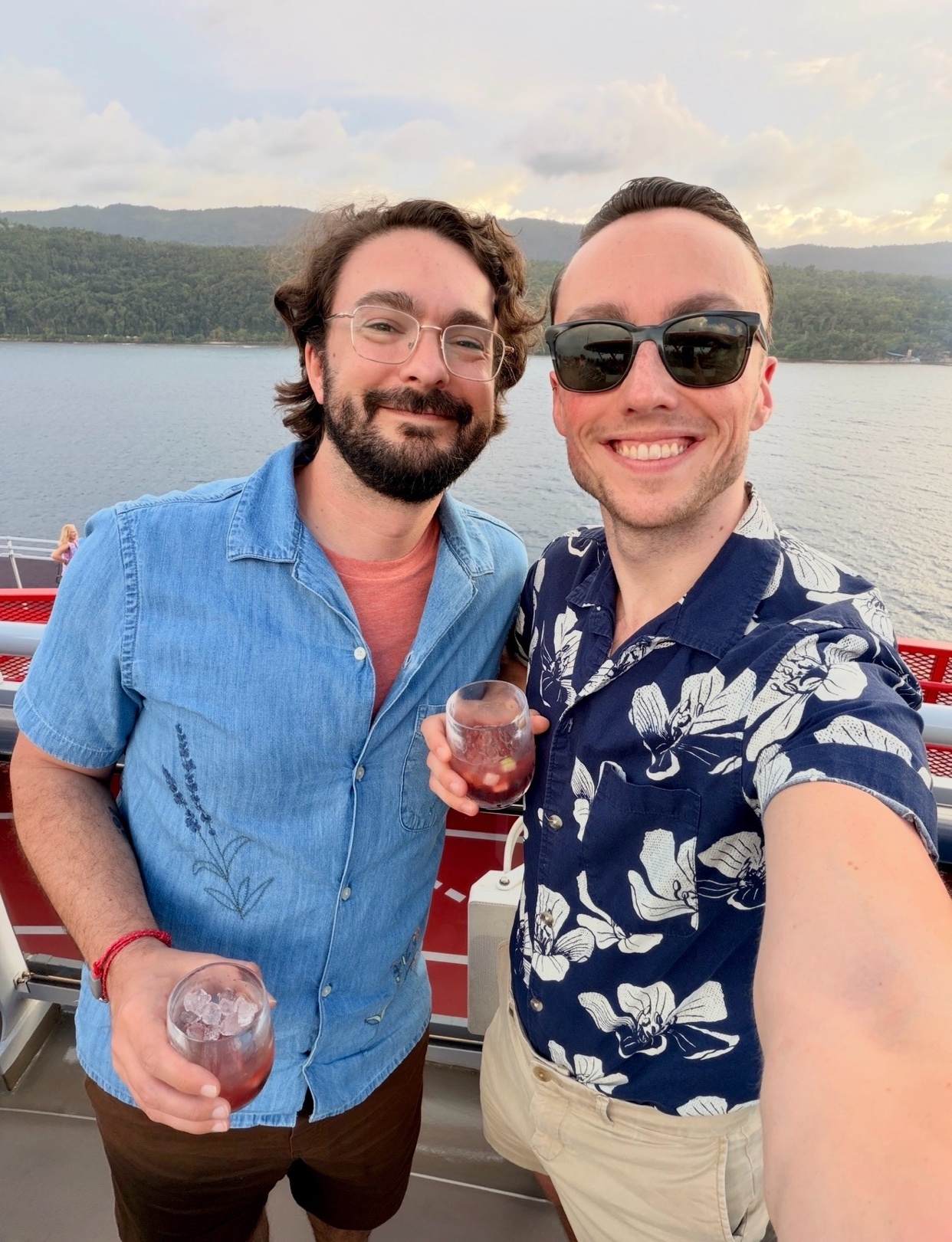 Two men smile for a selfie on a cruise ship deck, holding drinks. The man on the left wears glasses and a blue short-sleeved shirt, while the man on the right wears sunglasses and a floral print shirt.