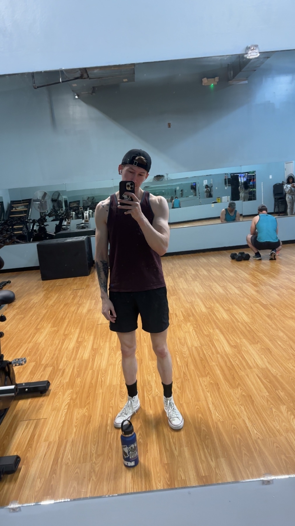 A man takes a mirror selfie in a gym, wearing a tank top, shorts, and a baseball cap. He is standing next to a water bottle with a tattoo visible on his arm.