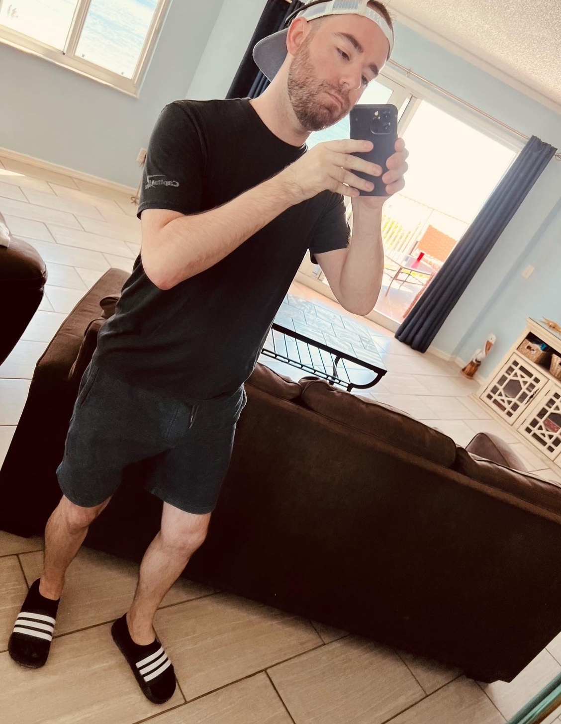 A man takes a selfie in a room while wearing a black t-shirt, dark shorts, and a baseball cap. He is also wearing black flip-flops with white stripes.