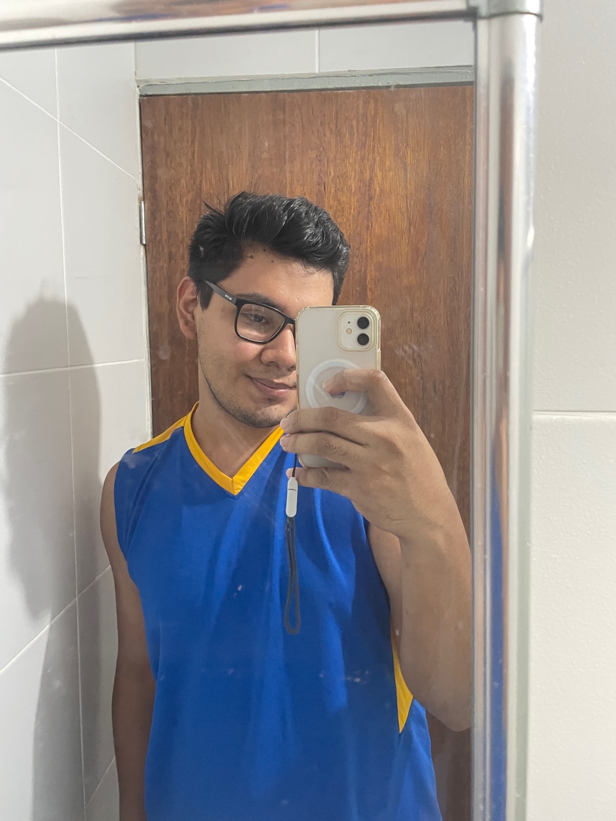 A man with dark hair and glasses takes a selfie in a bathroom mirror. He is wearing a blue sleeveless shirt with yellow trim and smiling at the camera.