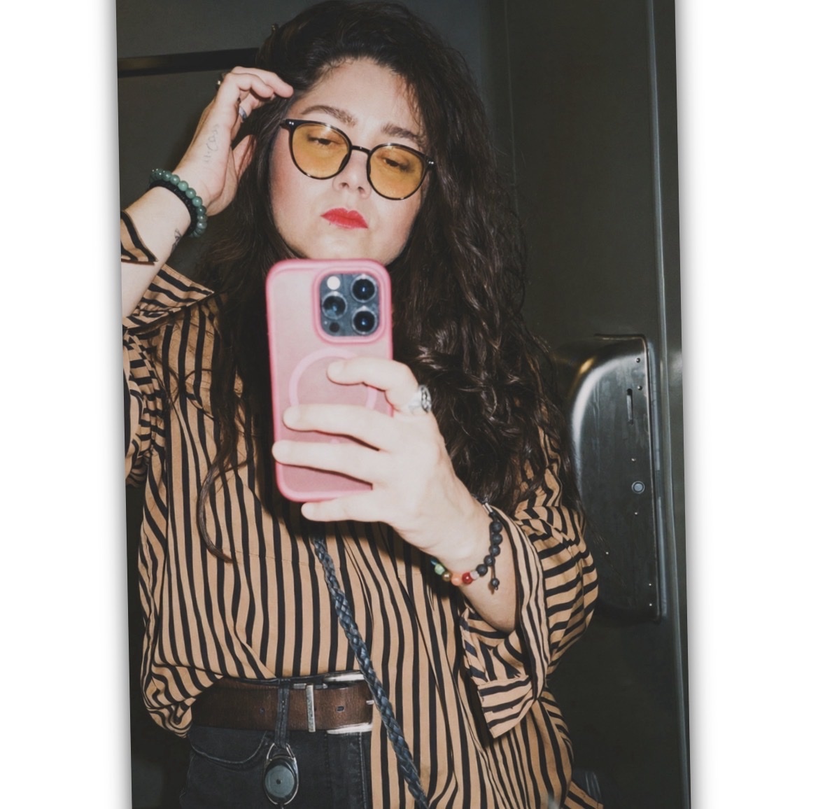 A woman with curly dark hair and glasses takes a selfie in a mirror. She is wearing a brown and black striped button-down shirt and red lipstick.
