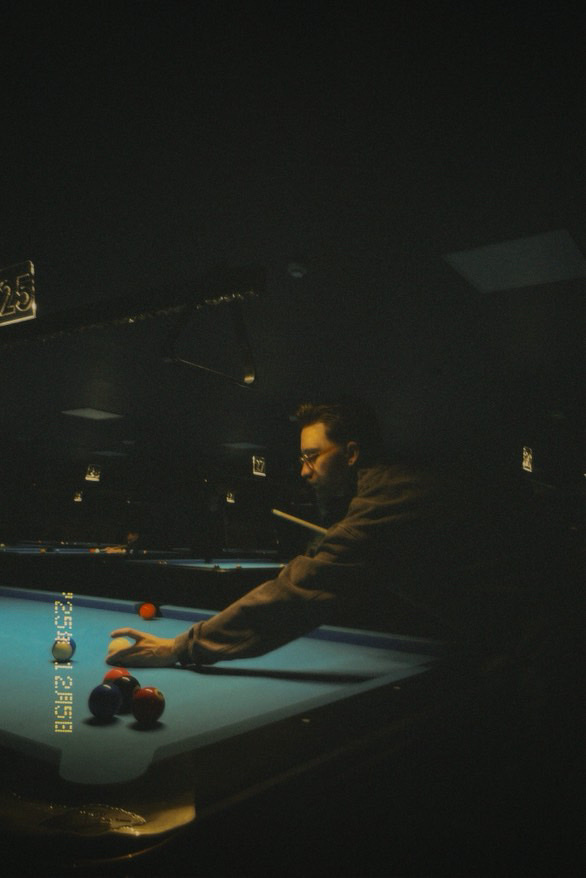 A man plays pool in a dimly lit room. He is focused on his shot, with the cue stick in hand.