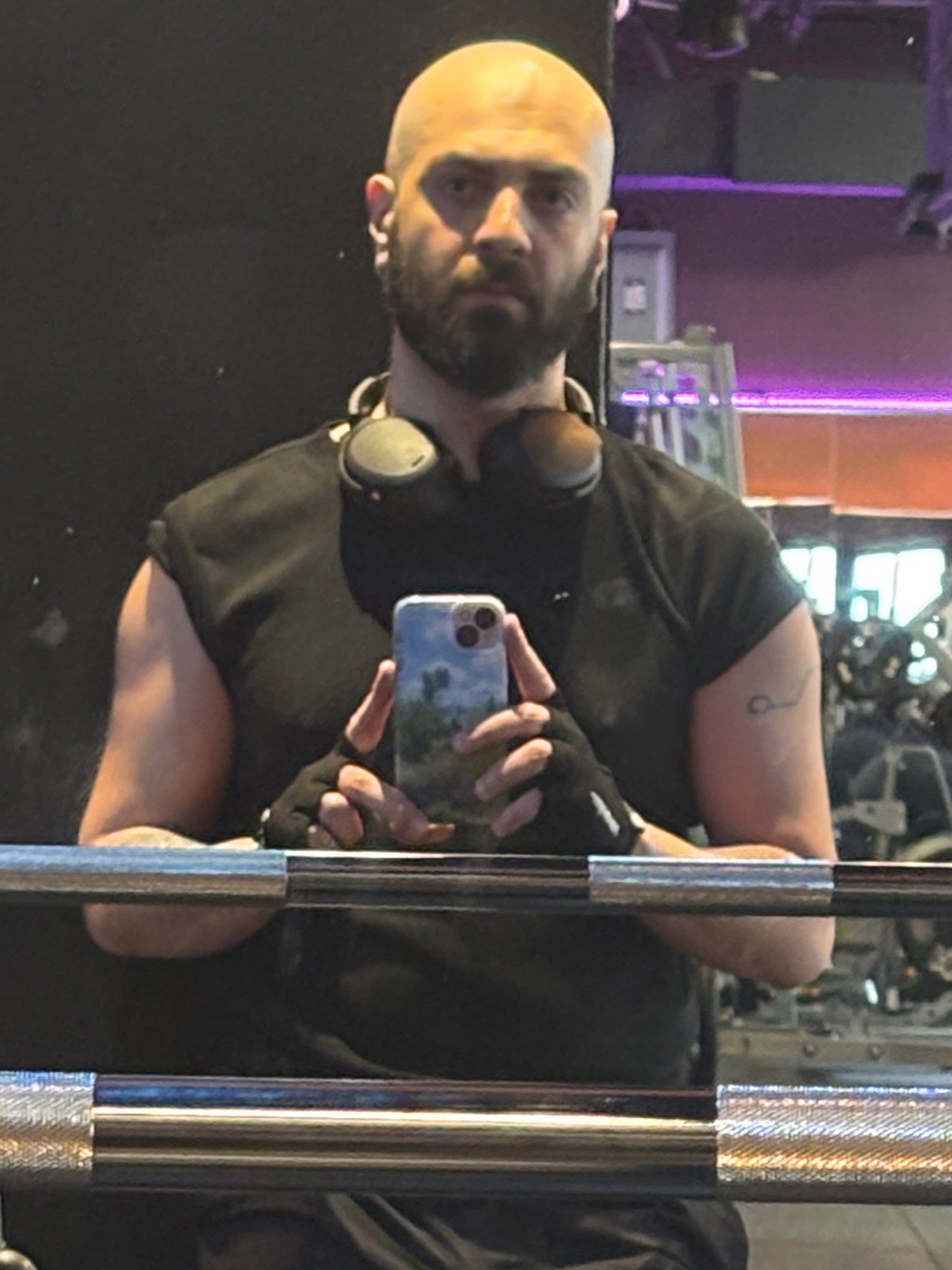 A bald man with a beard and headphones around his neck takes a mirror selfie in a gym. He is wearing a black t-shirt and fingerless gloves, and appears to be flexing his arm.