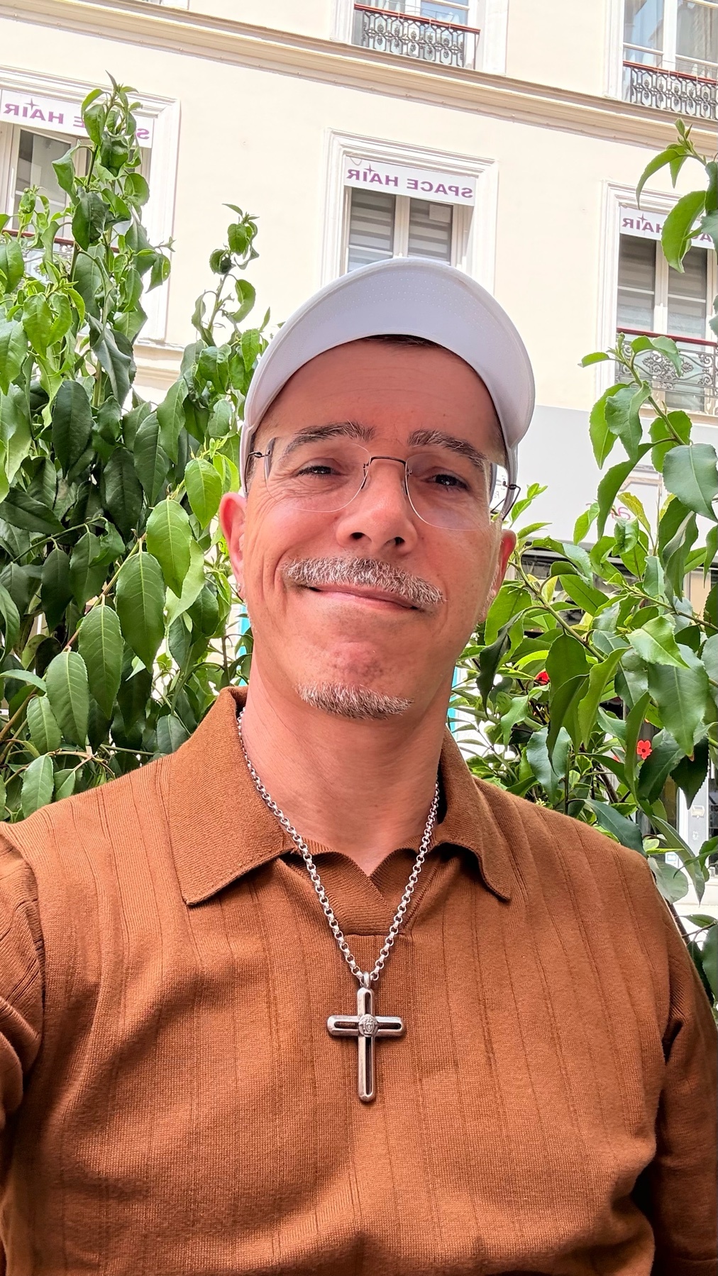 A man with glasses, stubble, and a mustache smiles at the camera while wearing a brown collared shirt, a silver cross necklace, and a white baseball cap. He is outdoors with green foliage and a building in the background.