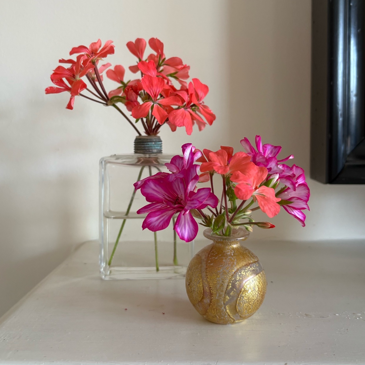 Two small vases hold vibrant pink and red flowers. The vase on the left is clear glass, while the one on the right is a small, ornate gold orb.
