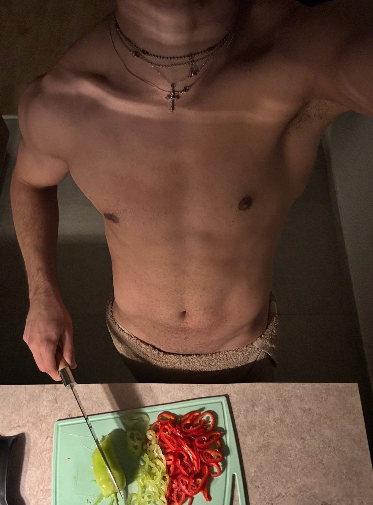 A shirtless, muscular man is preparing food in a kitchen. He is slicing bell peppers on a cutting board.