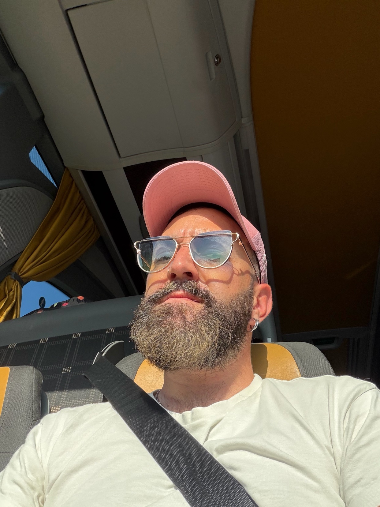 A man with a beard and sunglasses wears a pink baseball cap and a light-colored t-shirt. He is seated inside a vehicle, possibly a bus, with a seatbelt fastened across his chest.
