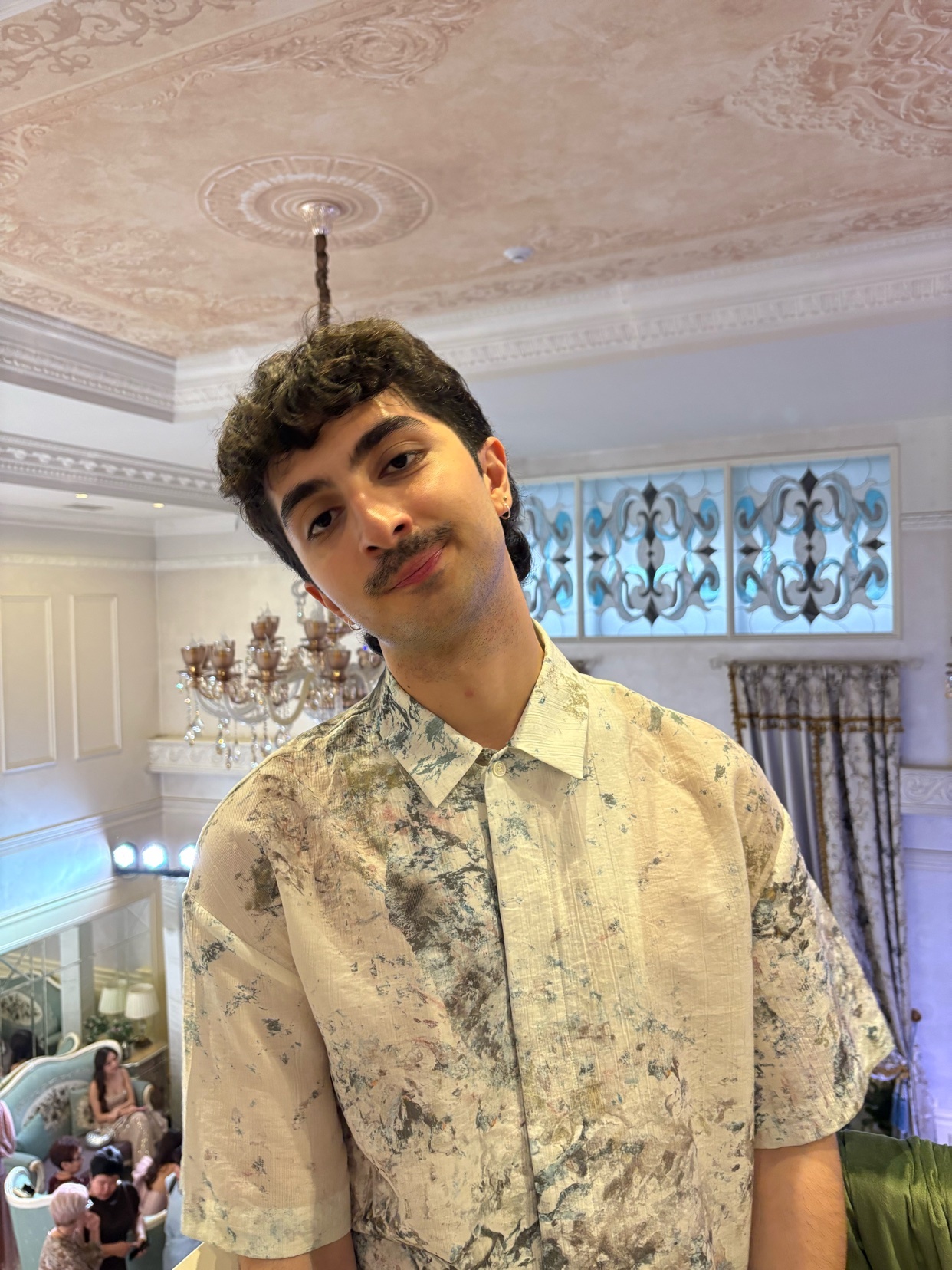 A man poses for a photo indoors. He is wearing a patterned shirt and has curly hair and a mustache.