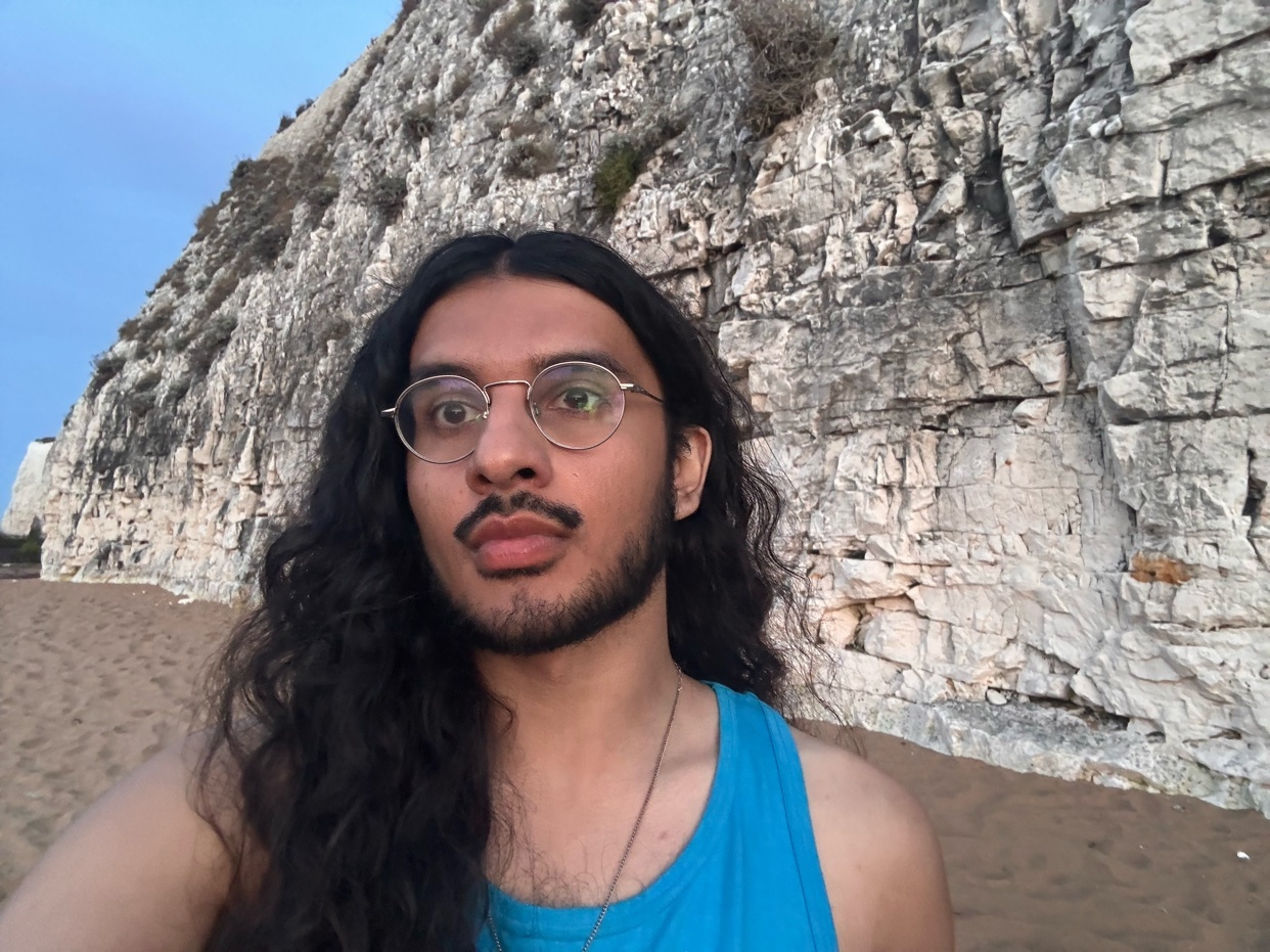 A man with long, dark, curly hair, a mustache, and a beard takes a selfie on a sandy beach with a large cliff face behind him. He is wearing round glasses and a blue tank top.