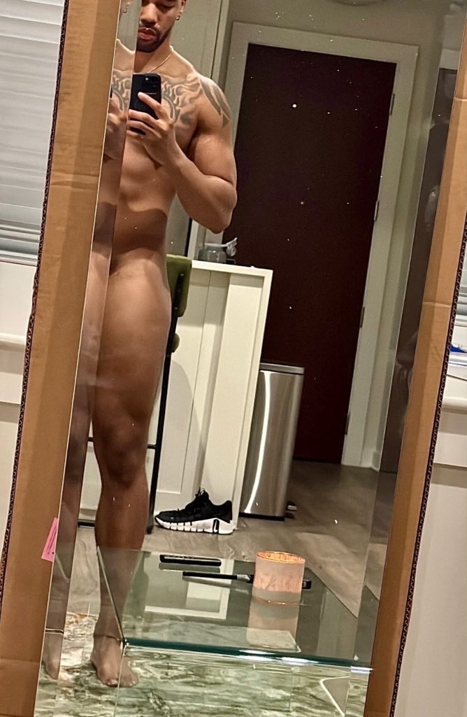 A shirtless man takes a mirror selfie, showcasing his muscular physique and tattoos. The photo highlights his physique and the reflection in the mirror.
