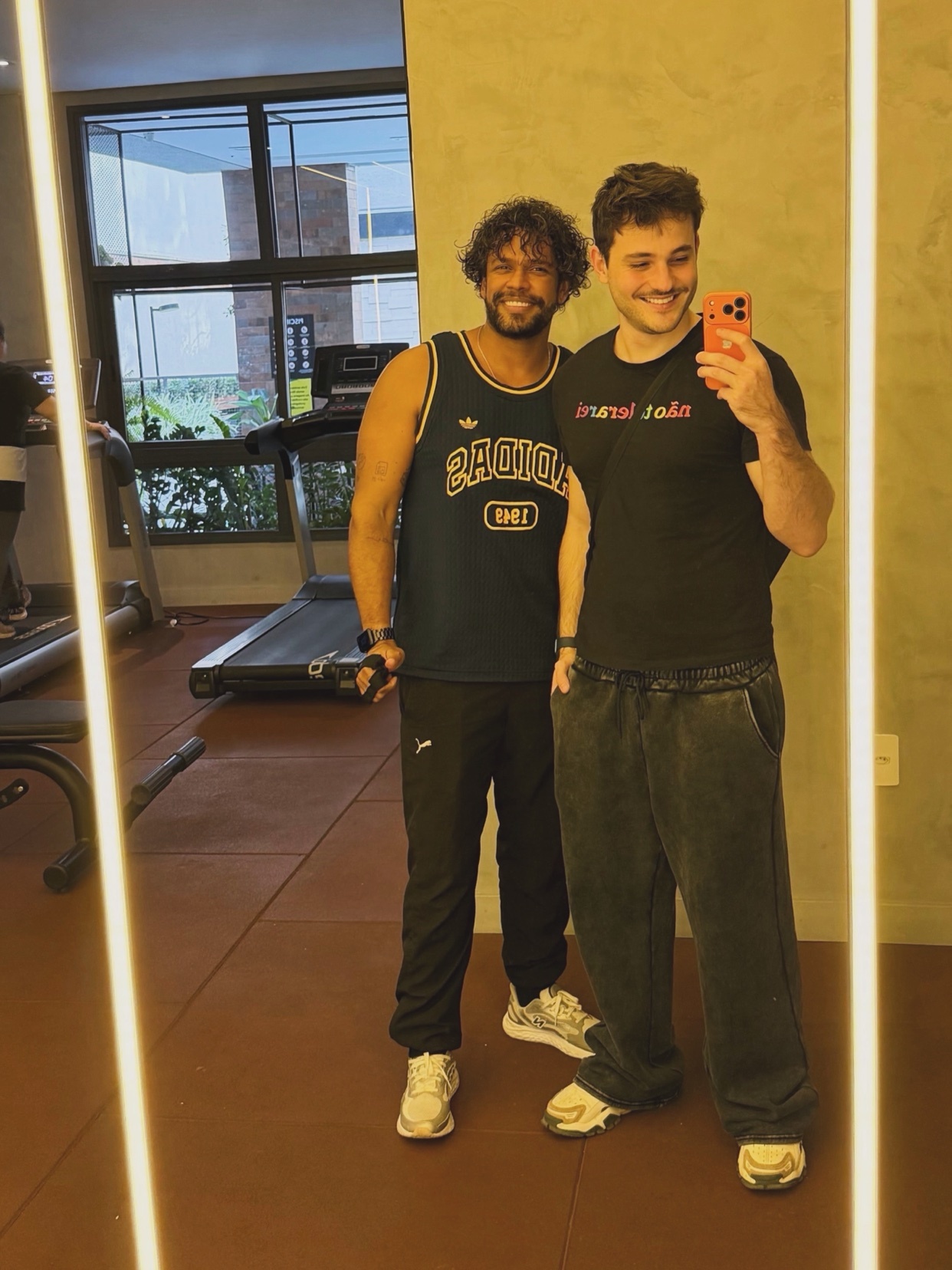 Two men pose for a mirror selfie in a gym. The man on the left wears a dark blue basketball jersey and black track pants, while the man on the right wears a black t-shirt and grey sweatpants. Both appear to be in good physical shape.