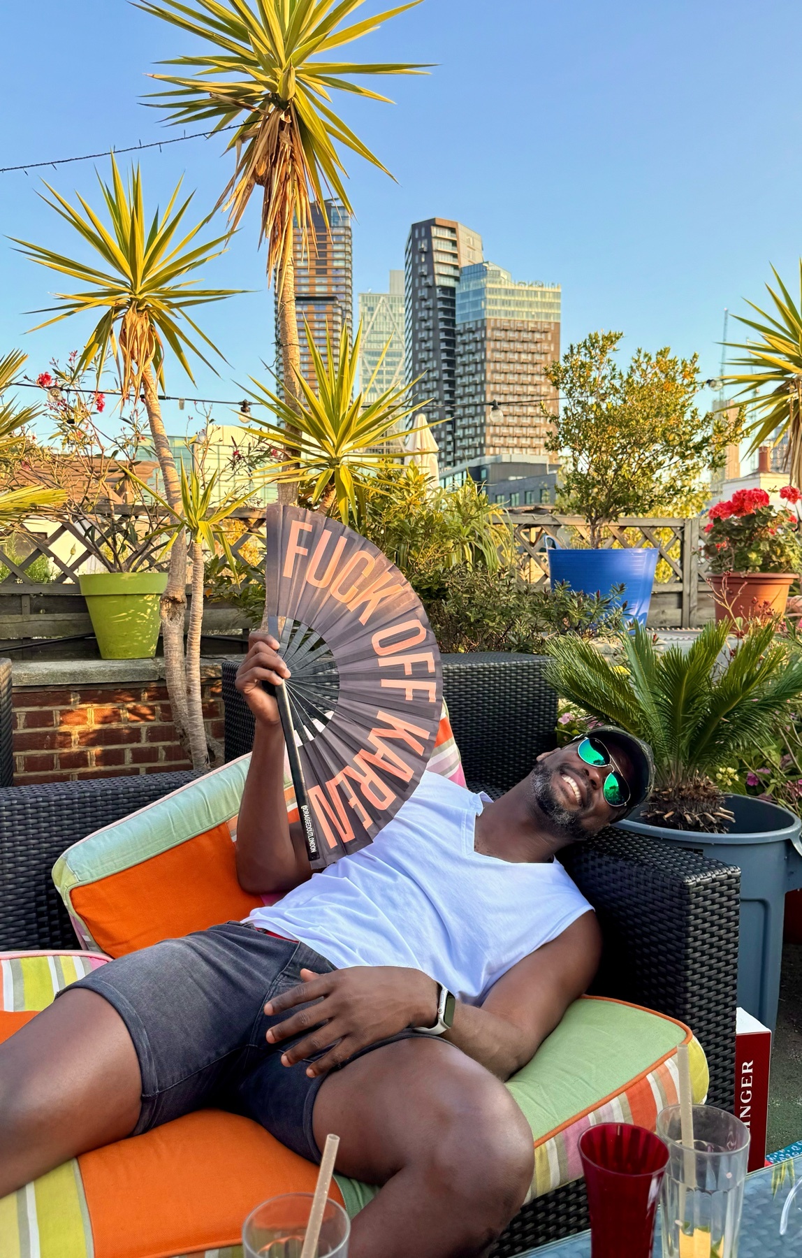 A man relaxes on a rooftop, holding a fan with the words "Fuck Off Karen" printed on it. The photo captures a sunny day with palm trees and city buildings in the background.