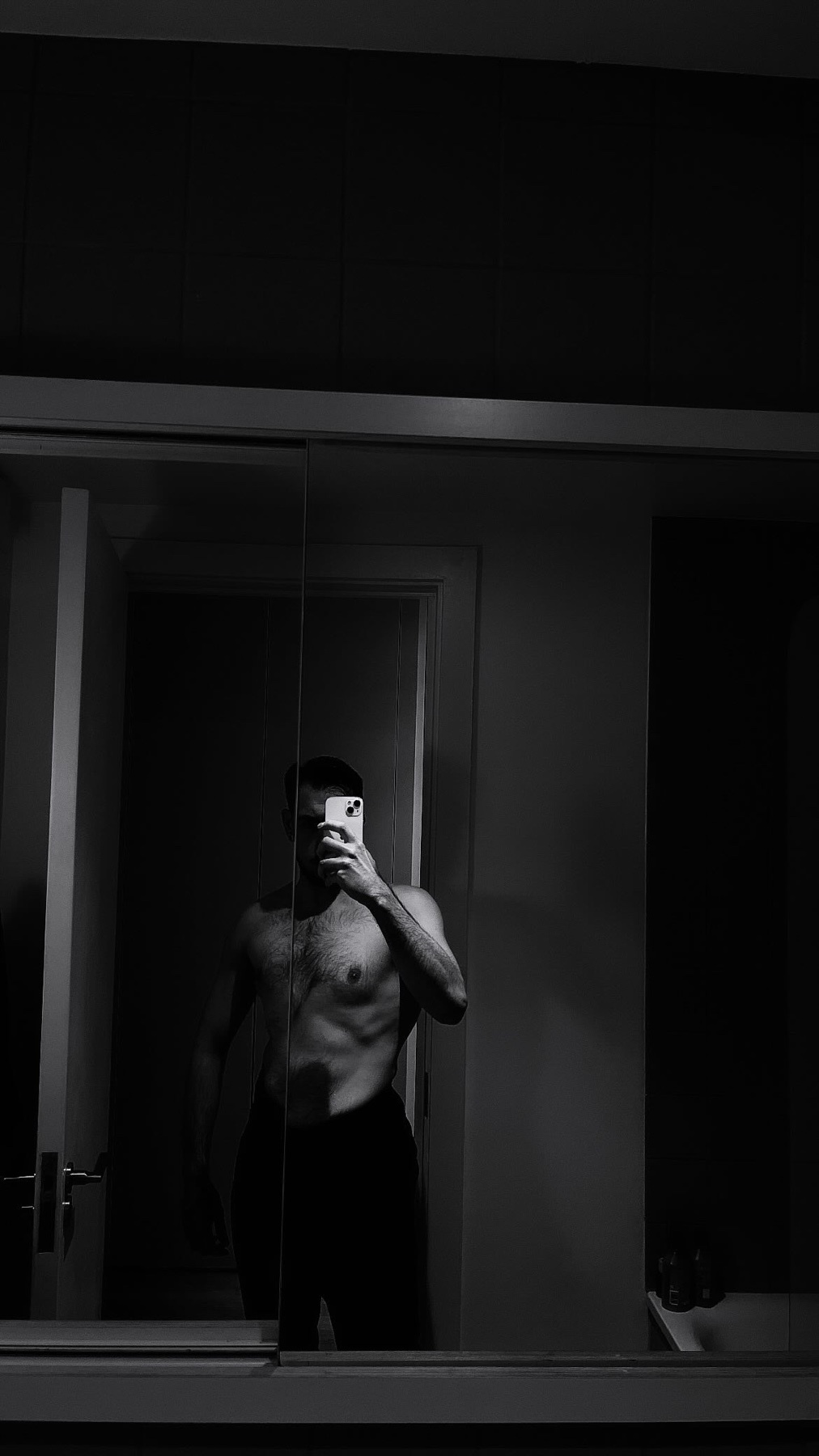 A man takes a mirror selfie, shirtless, showcasing his physique. The black and white photo emphasizes his muscular build.