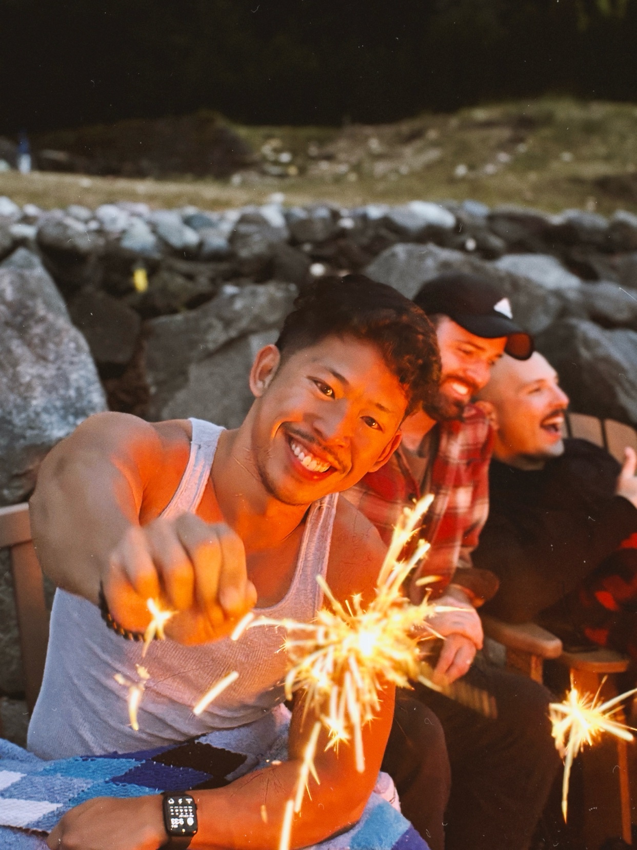 A man smiles while holding a sparkler, with two friends in the background. The photo captures a moment of joy and celebration.