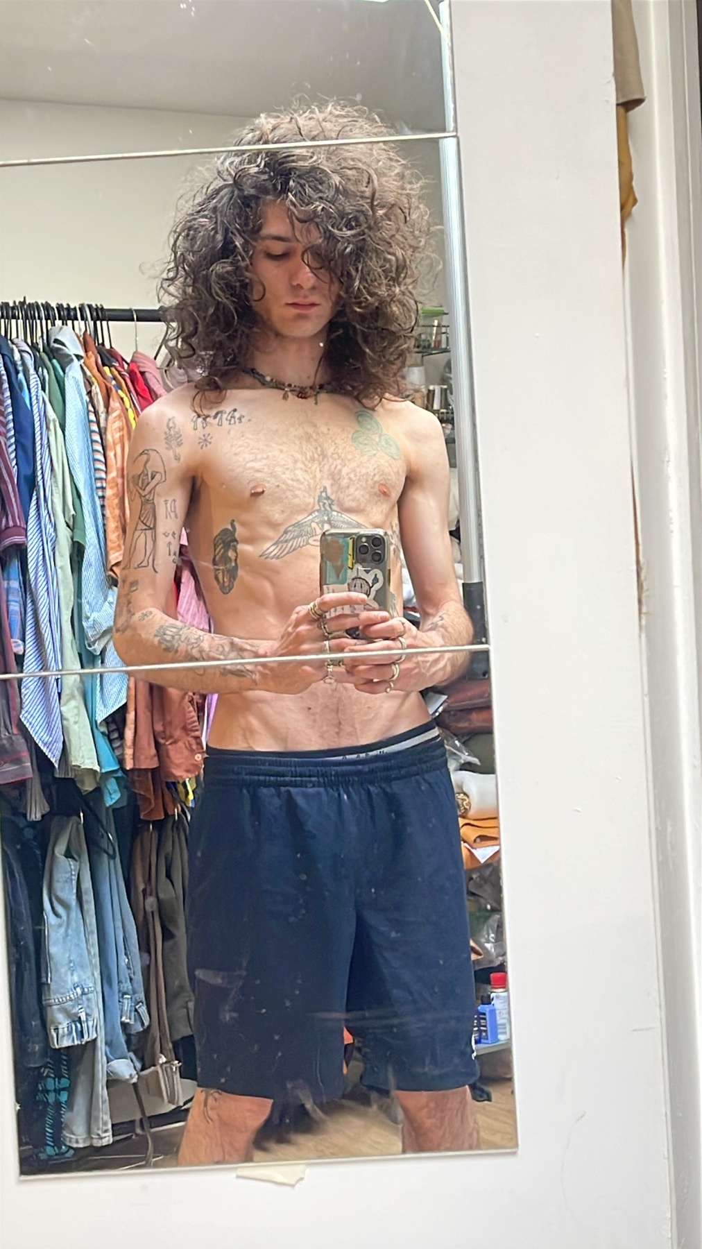 A shirtless man takes a mirror selfie. He has curly hair and tattoos.