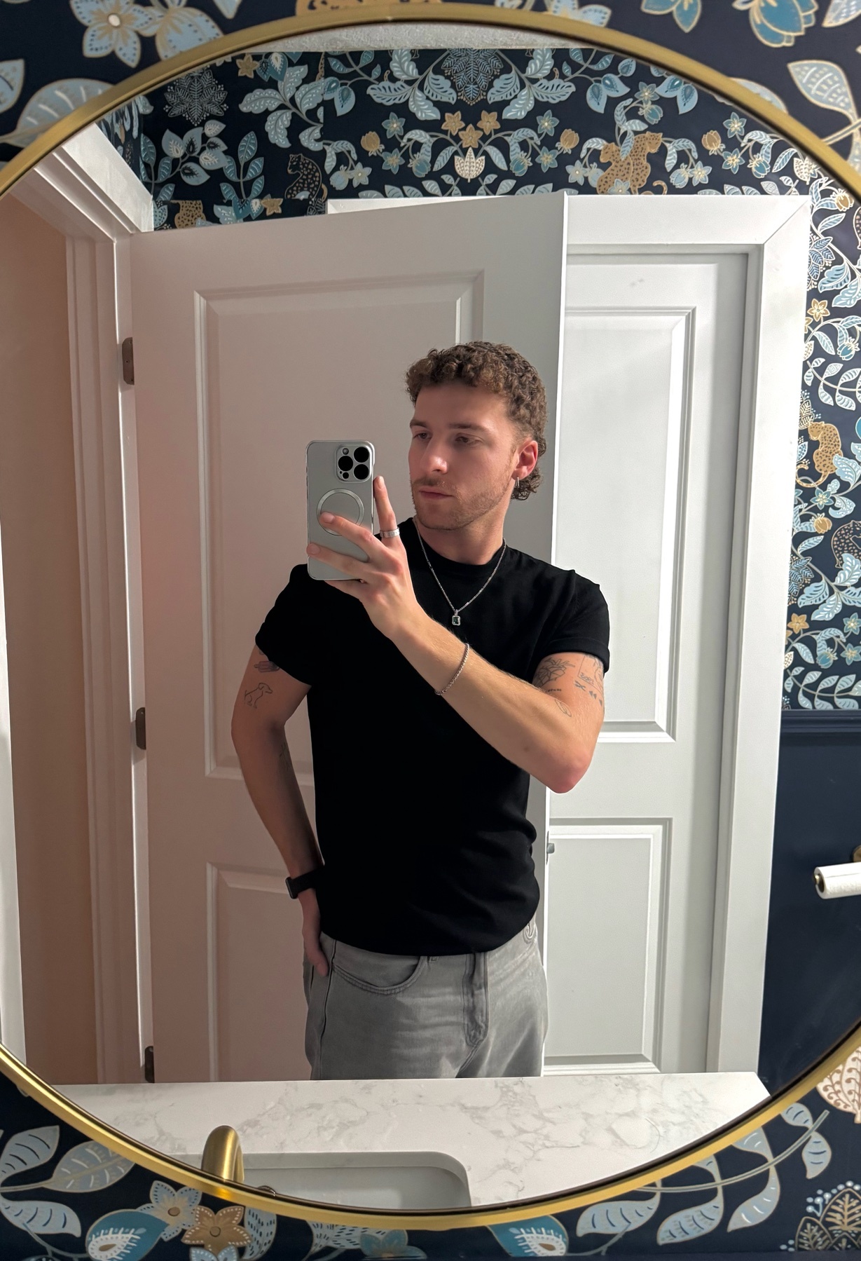 A man takes a mirror selfie in a bathroom. He is wearing a black t-shirt and grey pants.