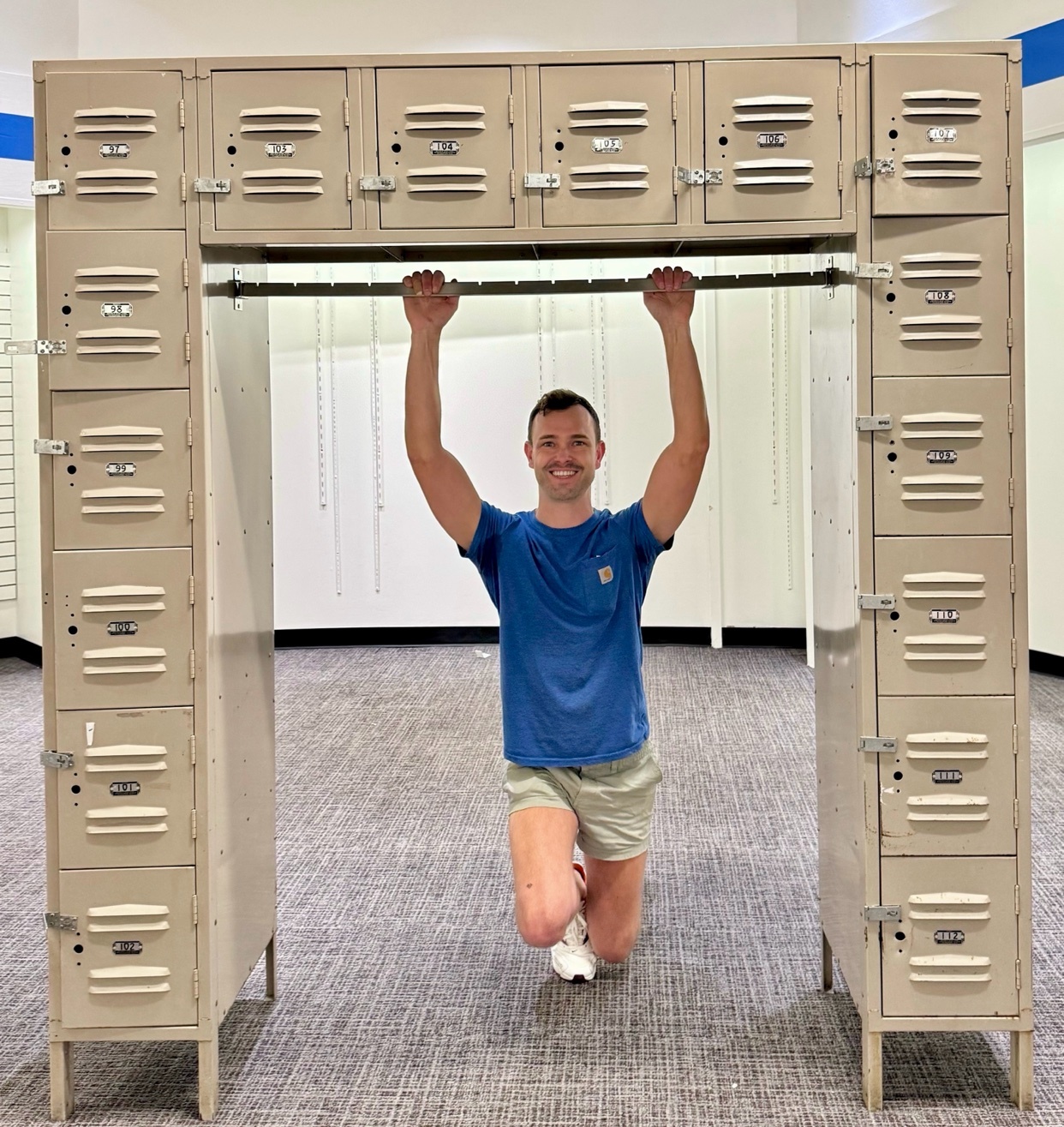 A man kneels and grips a pull-up bar mounted between two sets of lockers. He smiles at the camera, wearing a blue t-shirt and light-colored shorts.