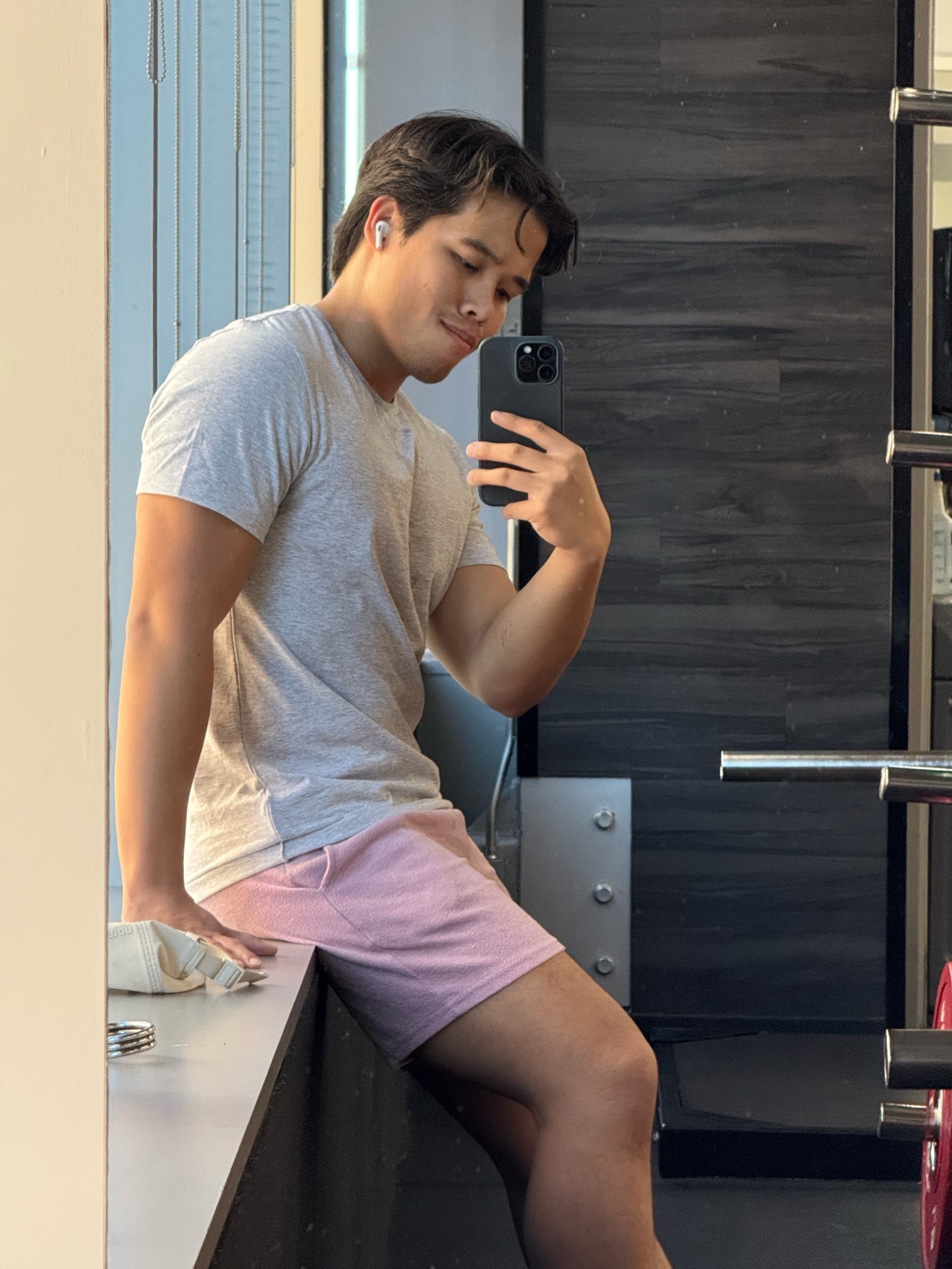 A man takes a mirror selfie in a gym, showcasing his physique. He's wearing a t-shirt and shorts, with gym equipment visible in the background.