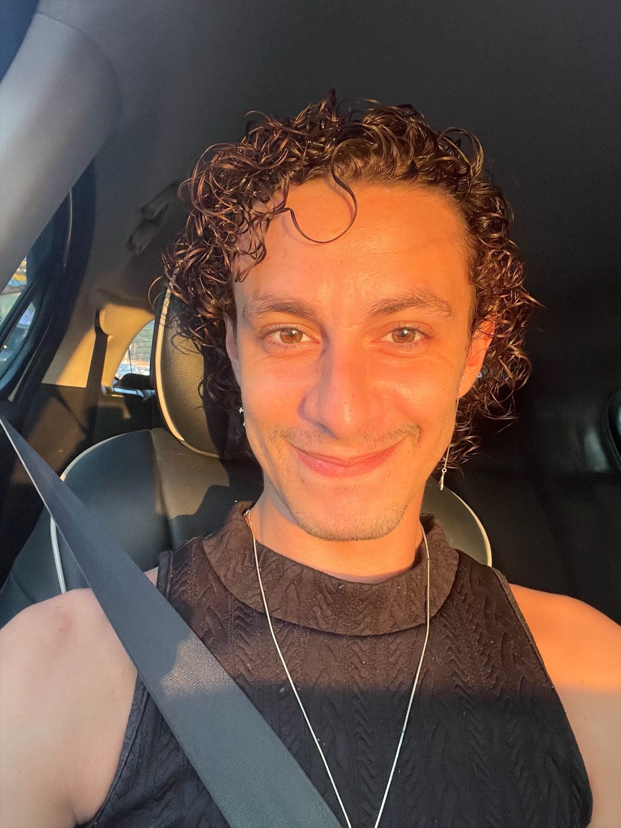 A man with curly dark hair and a warm smile takes a selfie in a car. The sunlight casts a warm glow on his face.