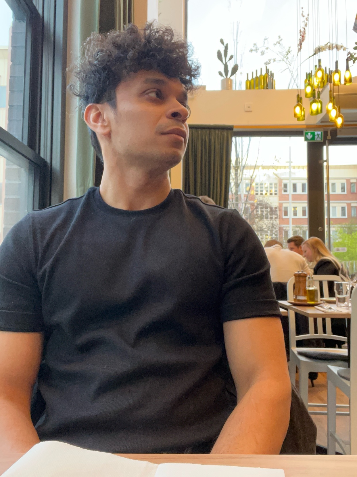 A man with curly dark hair sits at a table in a restaurant, looking off to the side. He is wearing a black t-shirt.
